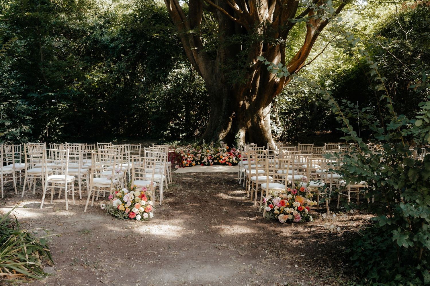Outdoor wedding ceremony setup with white chairs arranged under shady trees.