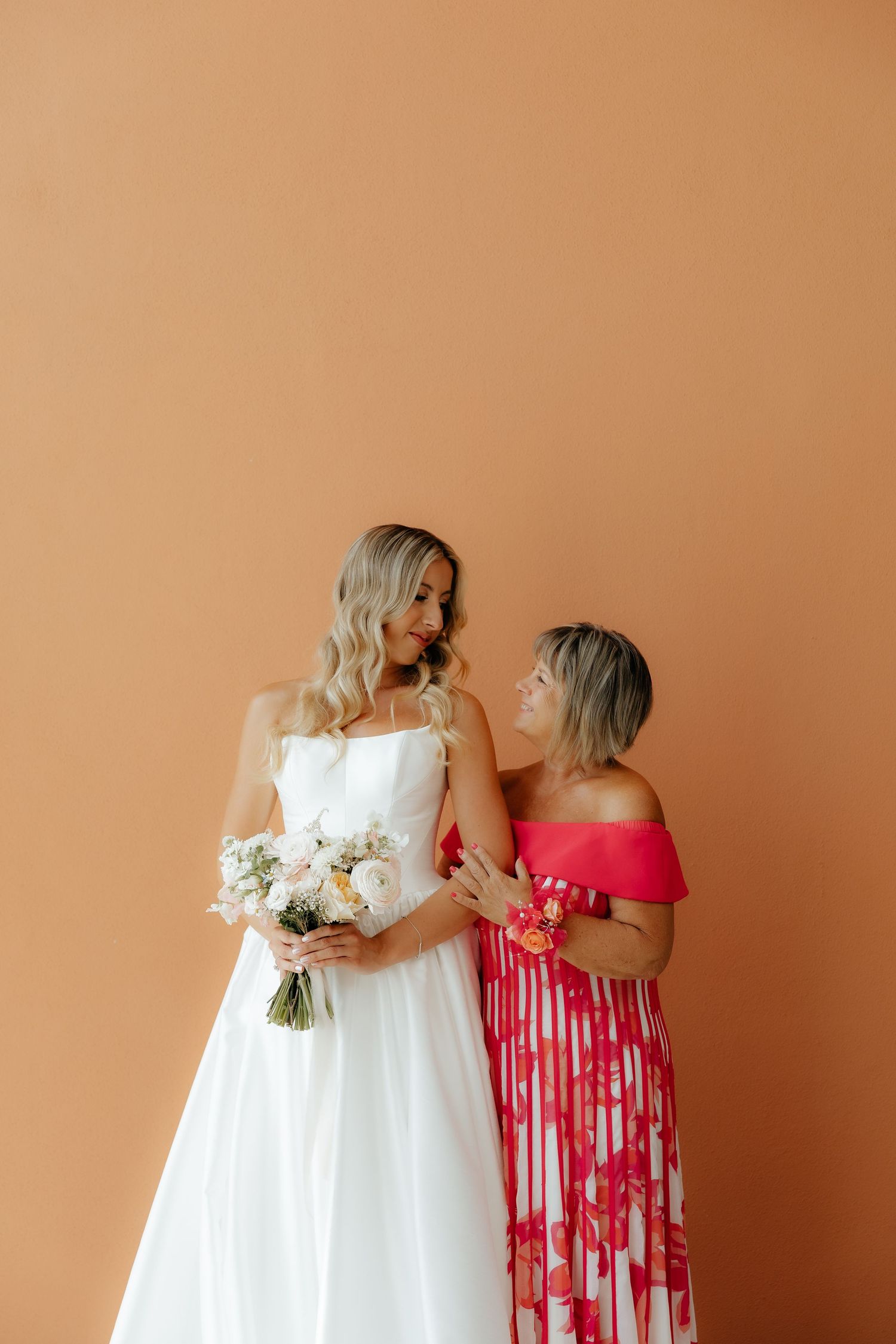 Two people share a tender moment against a peach wall, one in a white wedding gown and one in a red floral dress.