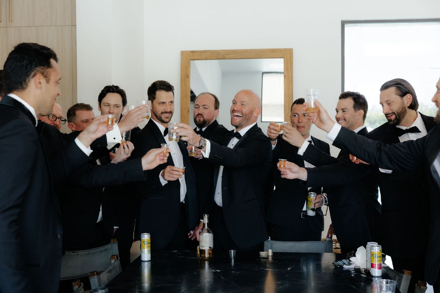 Groomsmen in black tuxedos toasting drinks together before wedding ceremony.