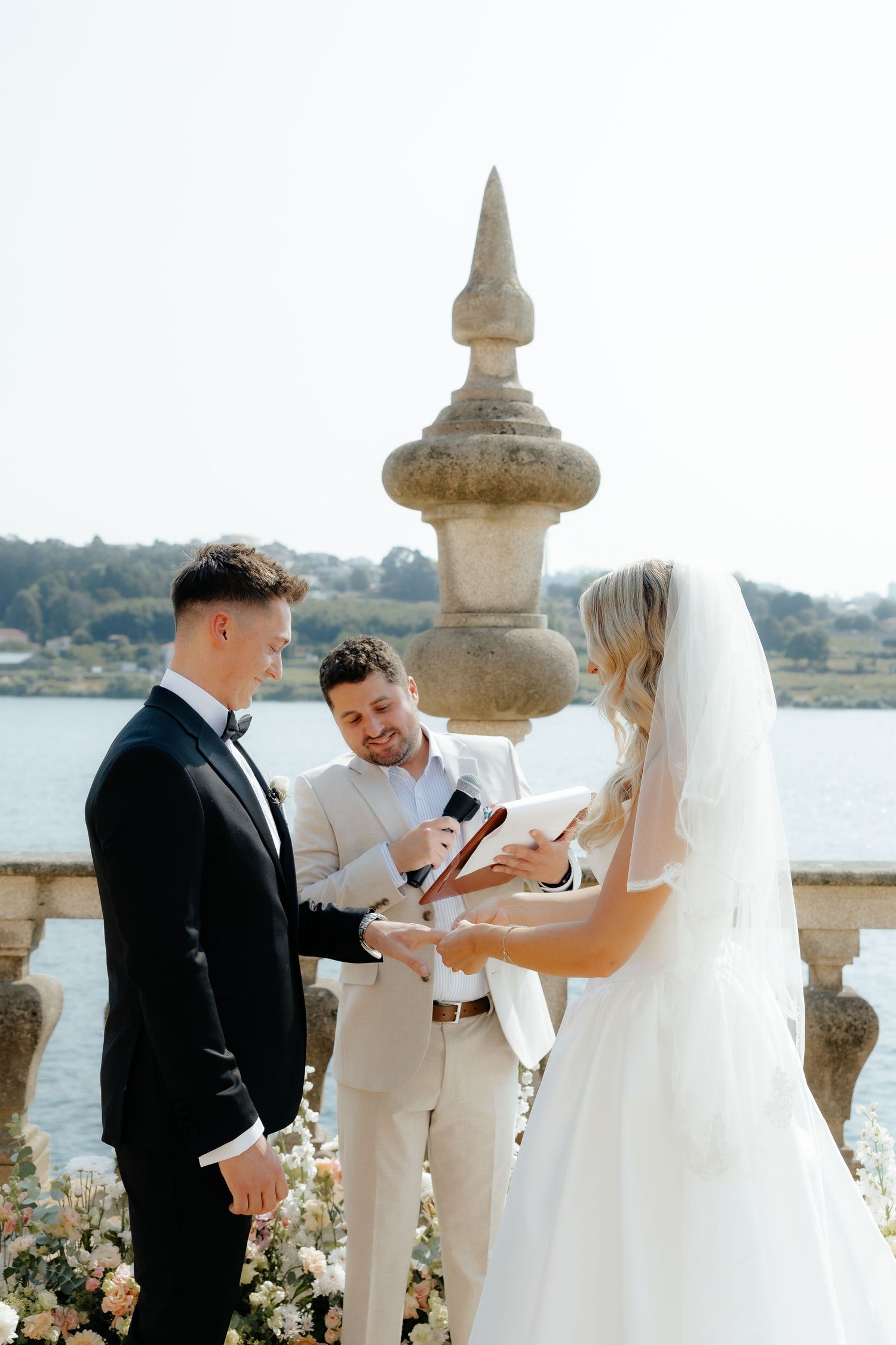 A wedding ceremony takes place on a stone terrace overlooking a lake with an ornate balustrade in the background.