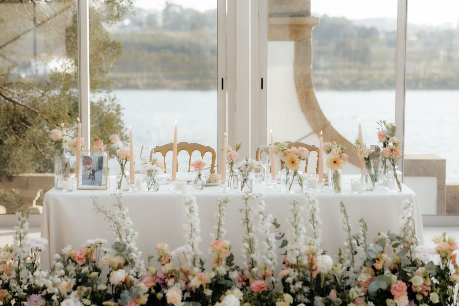 Elegant wedding reception table with floral arrangements and place settings overlooking a waterfront view through large windows.