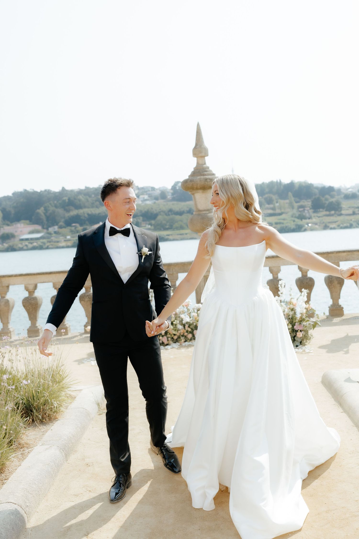 Smiling bride and groom walk hand in hand on scenic stone terrace overlooking river after wedding.
