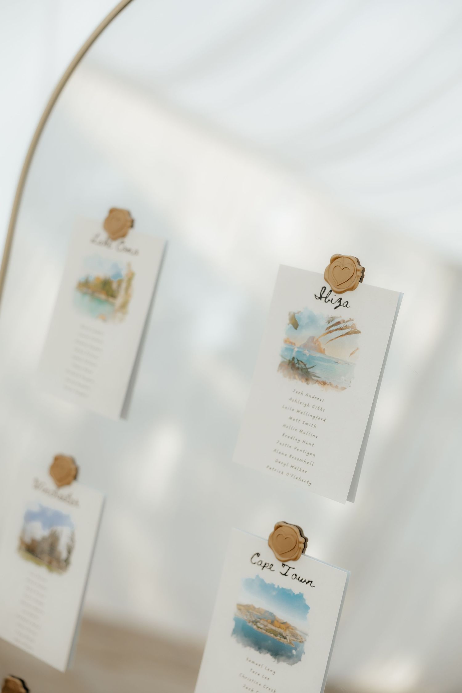 Close-up of watercolor destination wedding seating chart cards with gold wax seals on a mirror display.