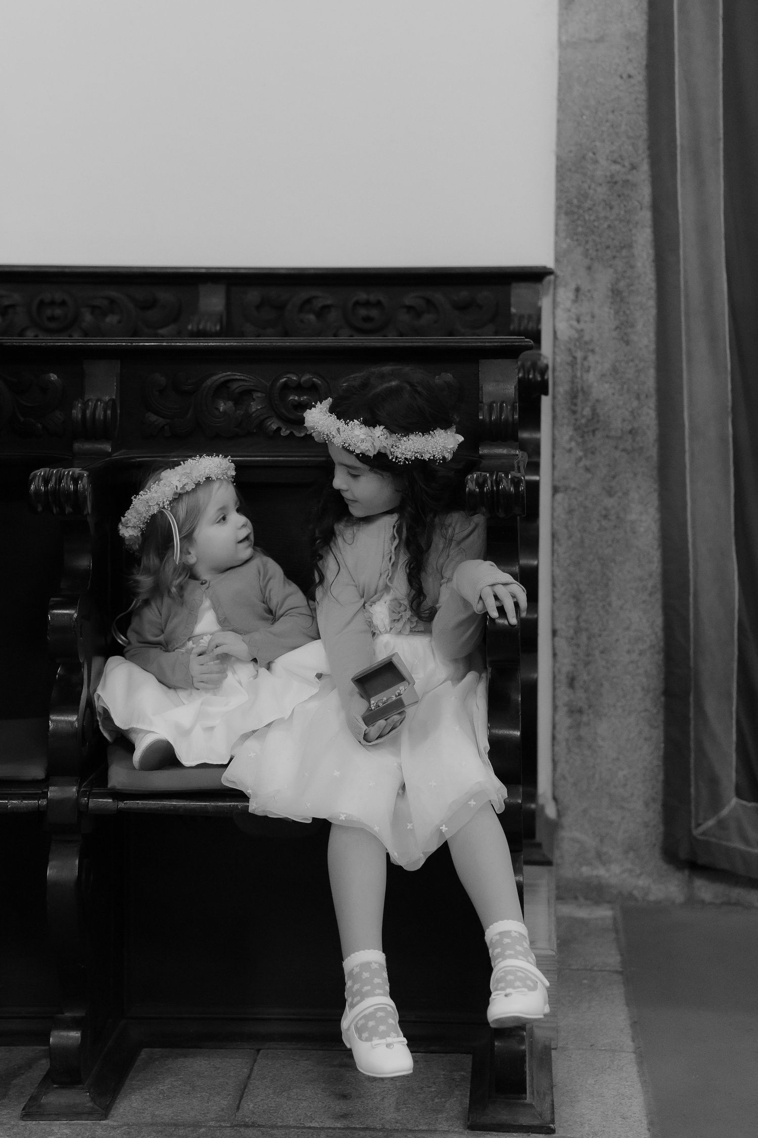 Two young flower girls in white dresses and floral crowns sit on church bench.