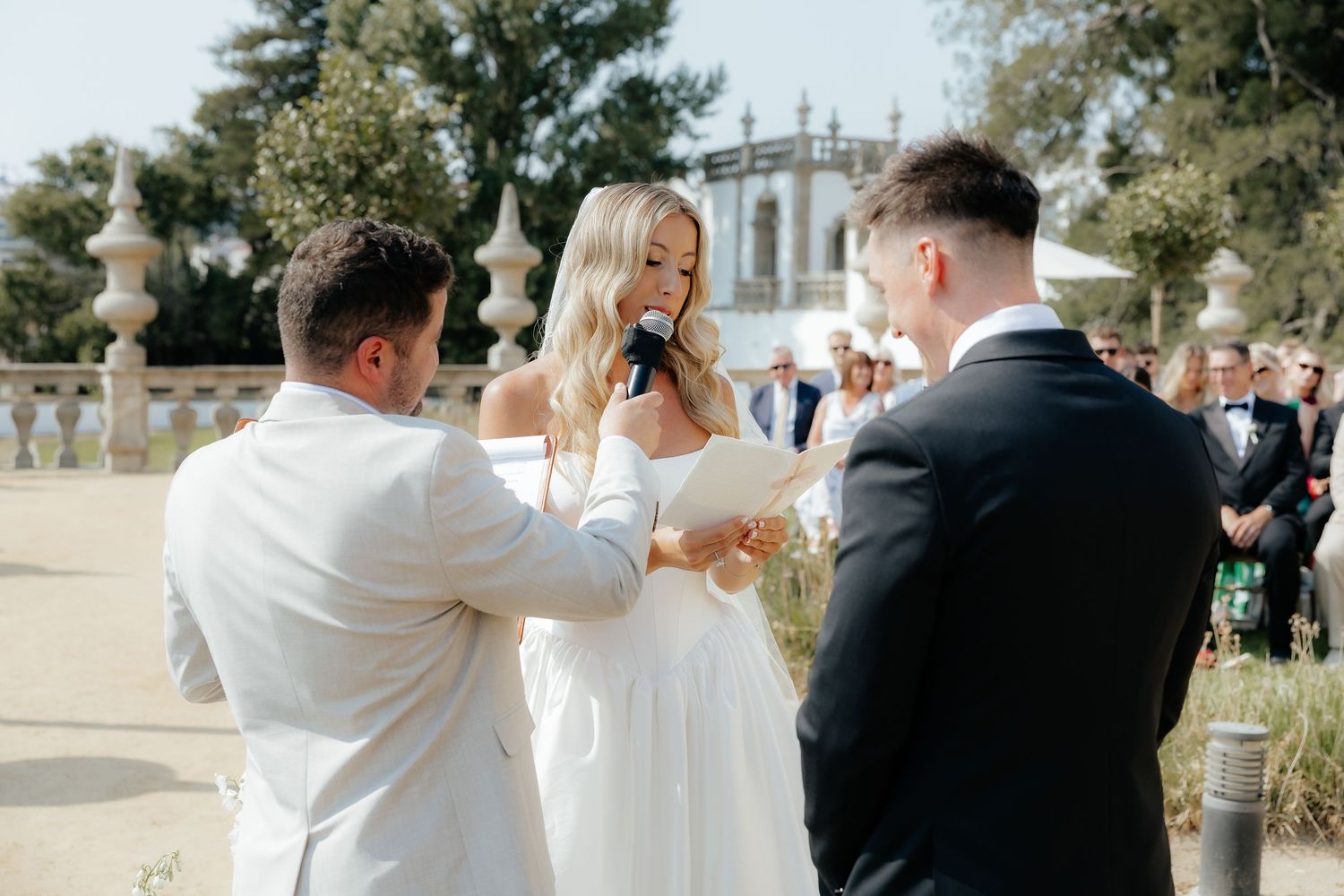 Blonde bride in white dress reads vows at outdoor wedding ceremony with groom and officiant present.