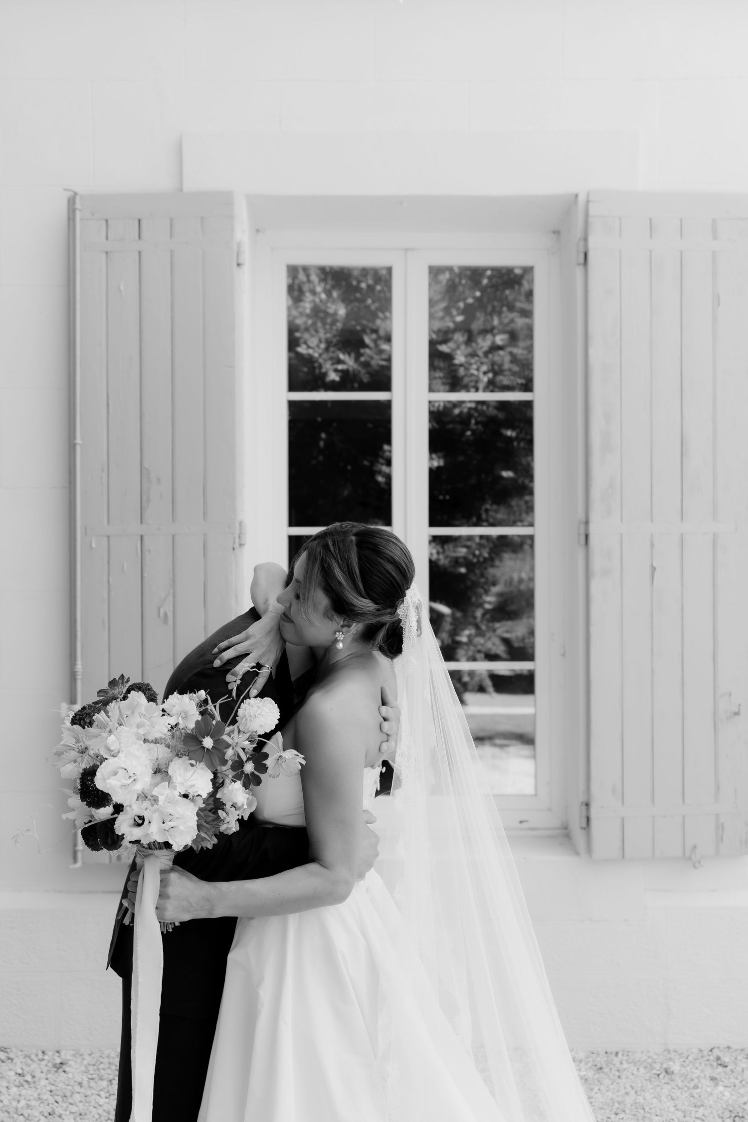 Black and white photo of bride with bouquet against white exterior with shuttered windows.