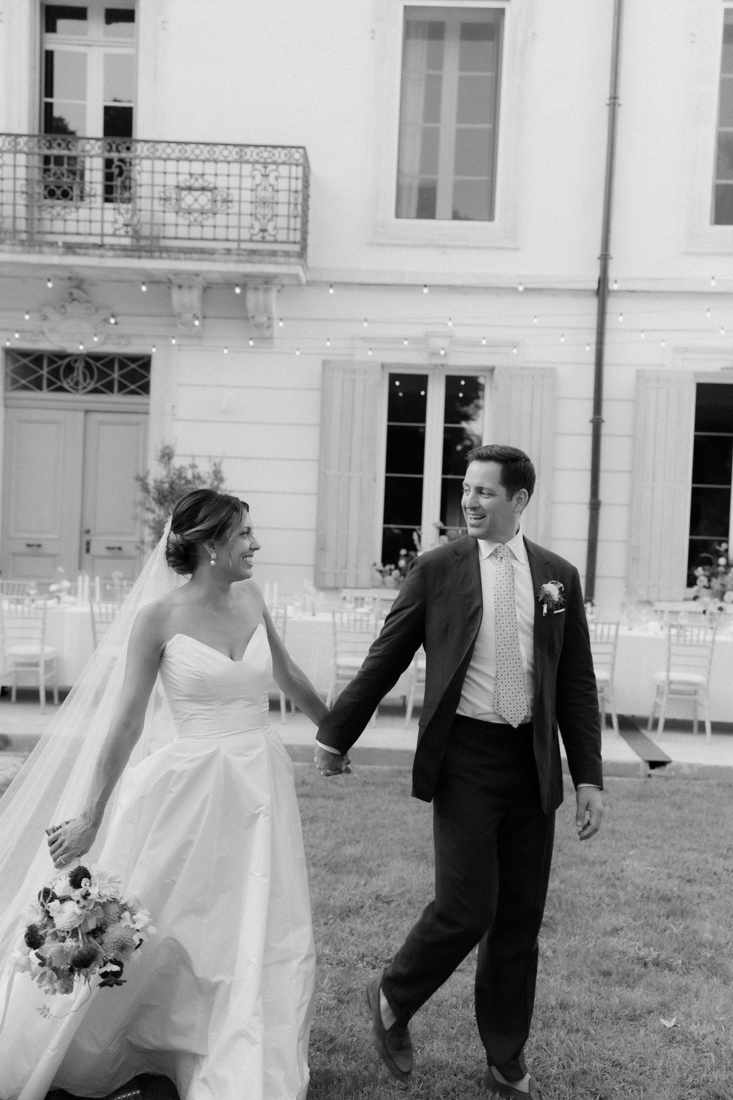 A black and white photo of newlyweds walking together on grass outside an elegant building.