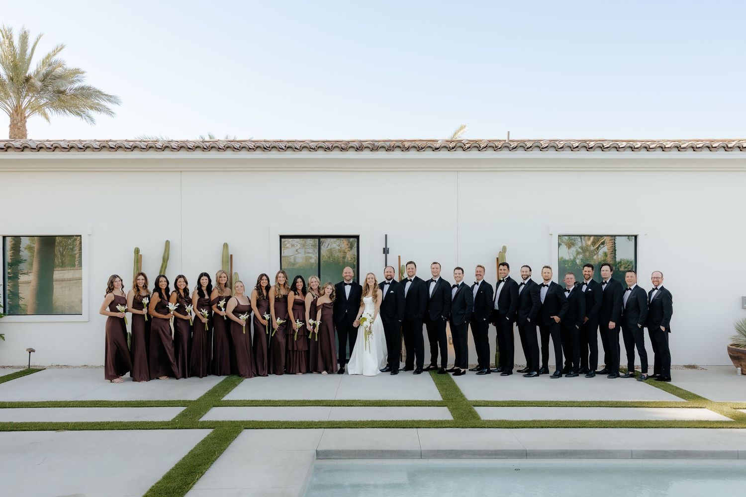 Wedding party poses together outdoors featuring bridesmaids in brown gowns and groomsmen in black tuxedos.