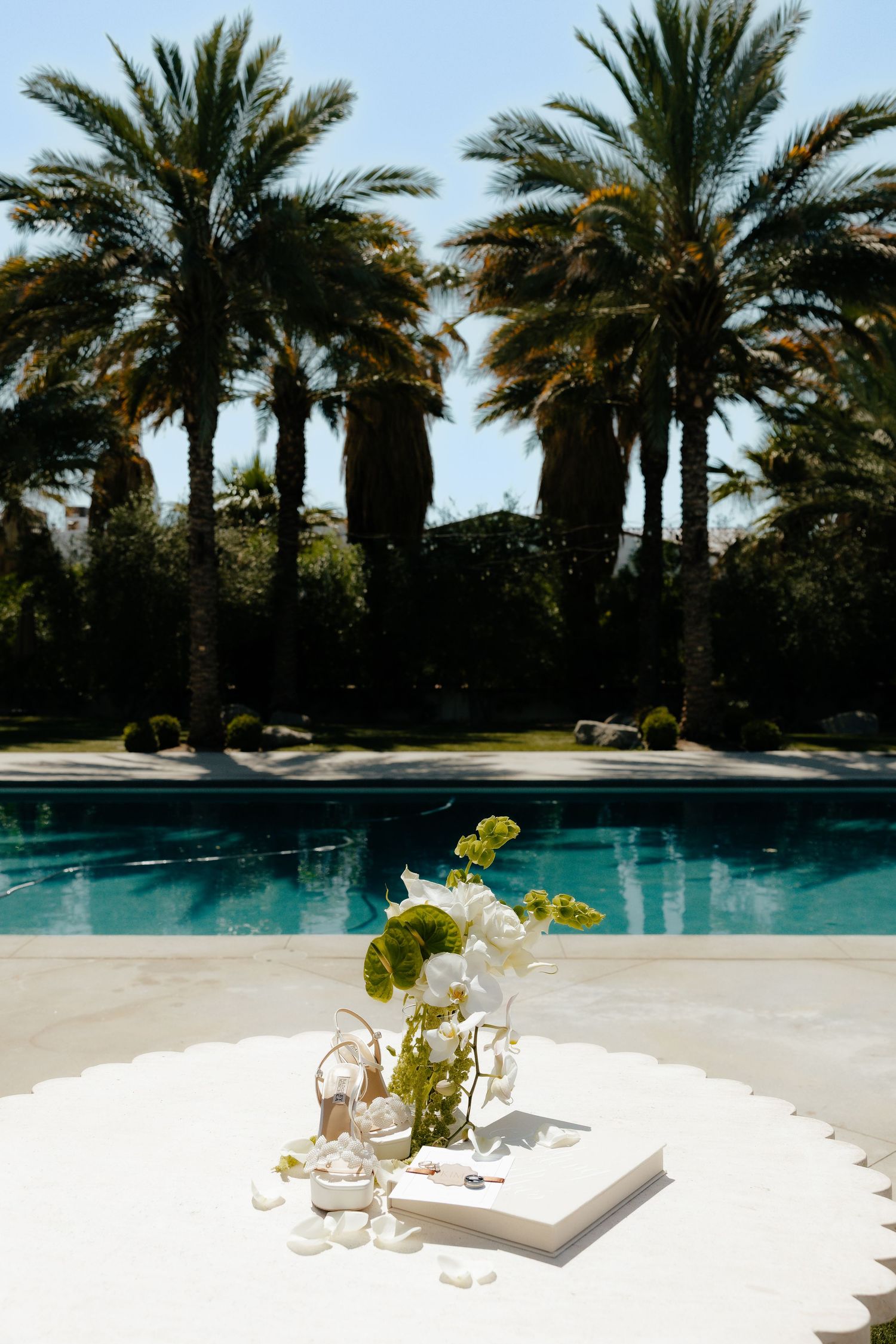 White floral arrangement and open book displayed on a poolside table with palm trees reflecting in blue water.