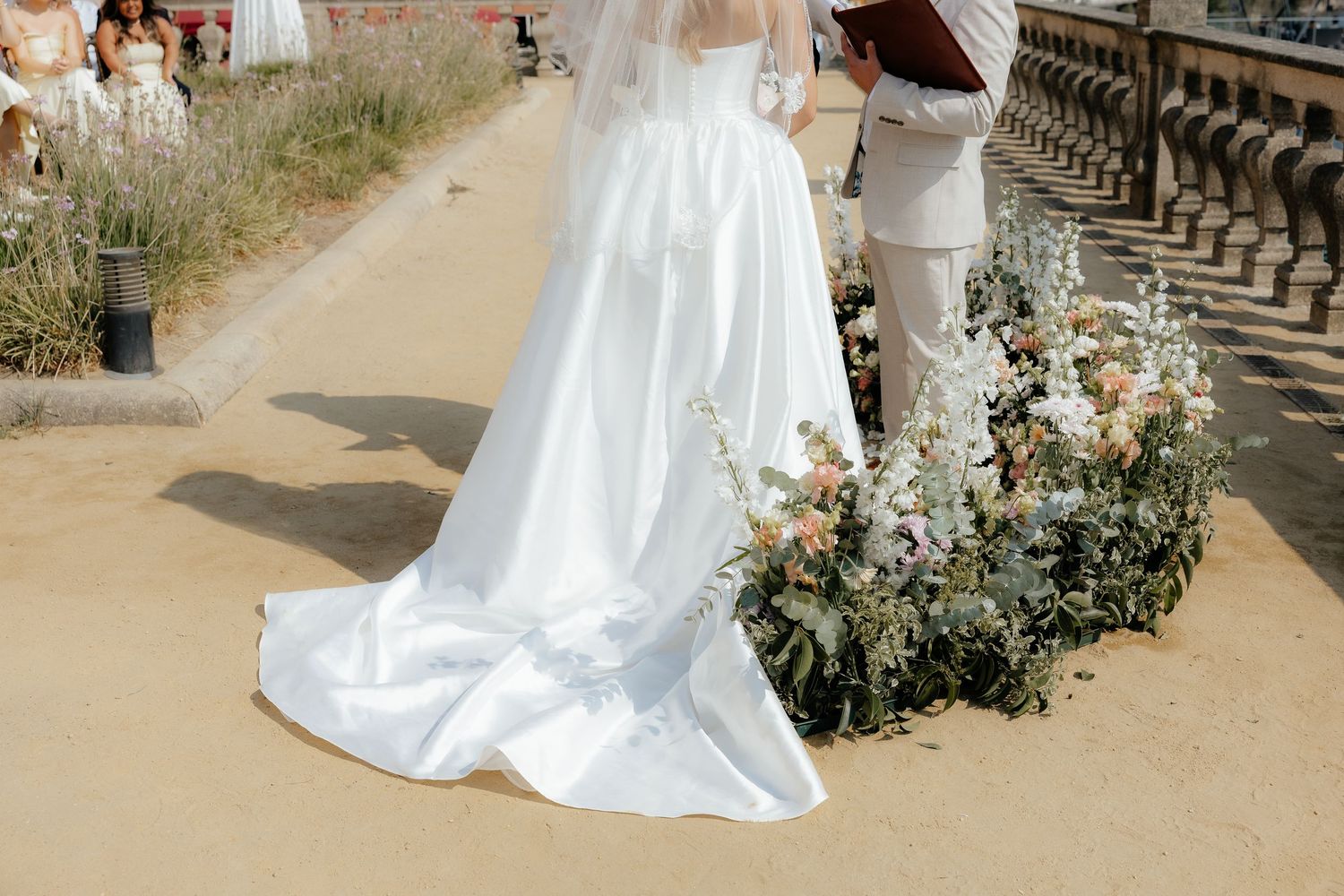 Bride in flowing white gown with long train stands beside floral arrangement during outdoor wedding ceremony.