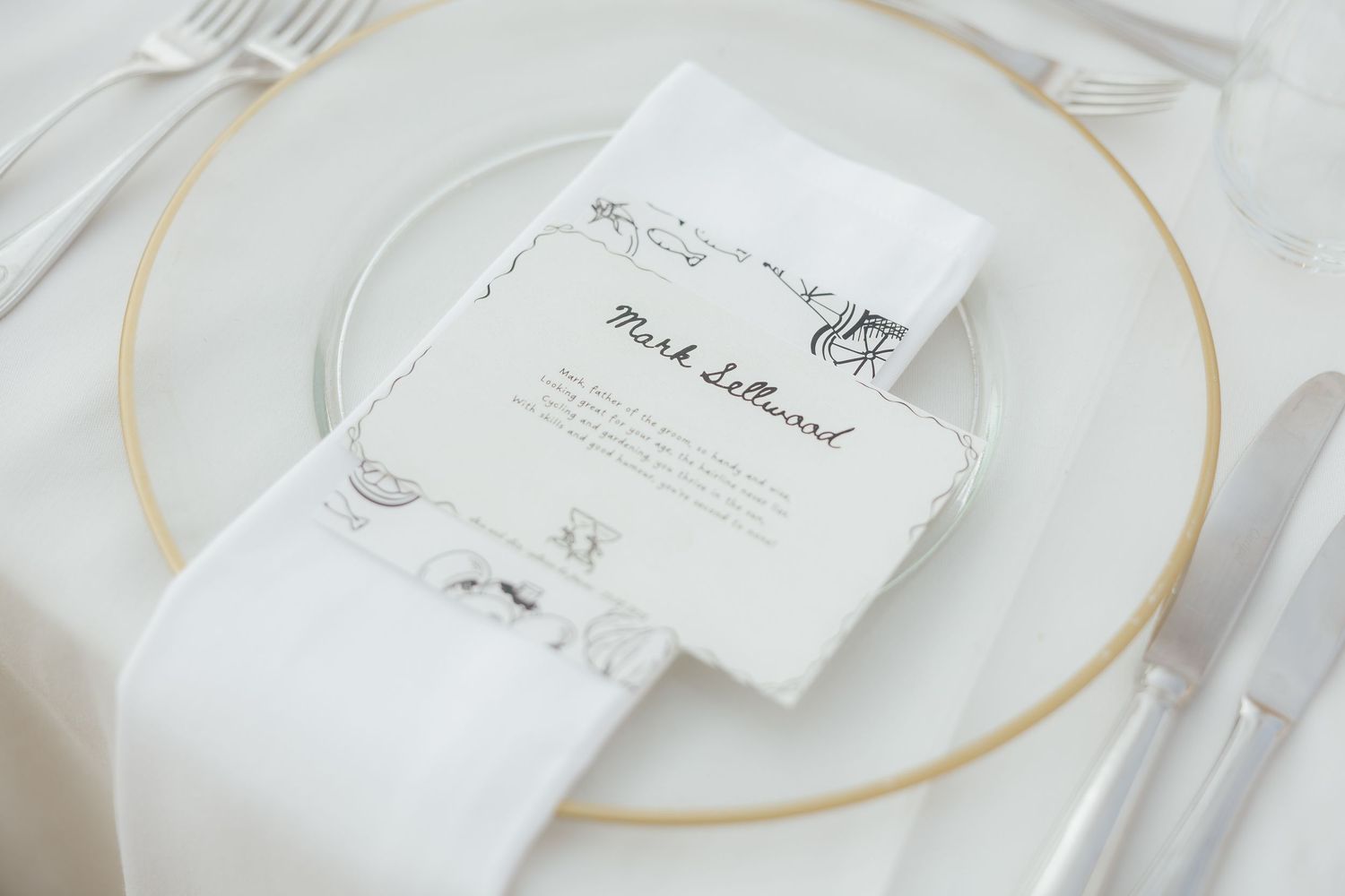 Minimalist wedding place setting with gold-rimmed charger plate, white napkin, and handwritten menu card.