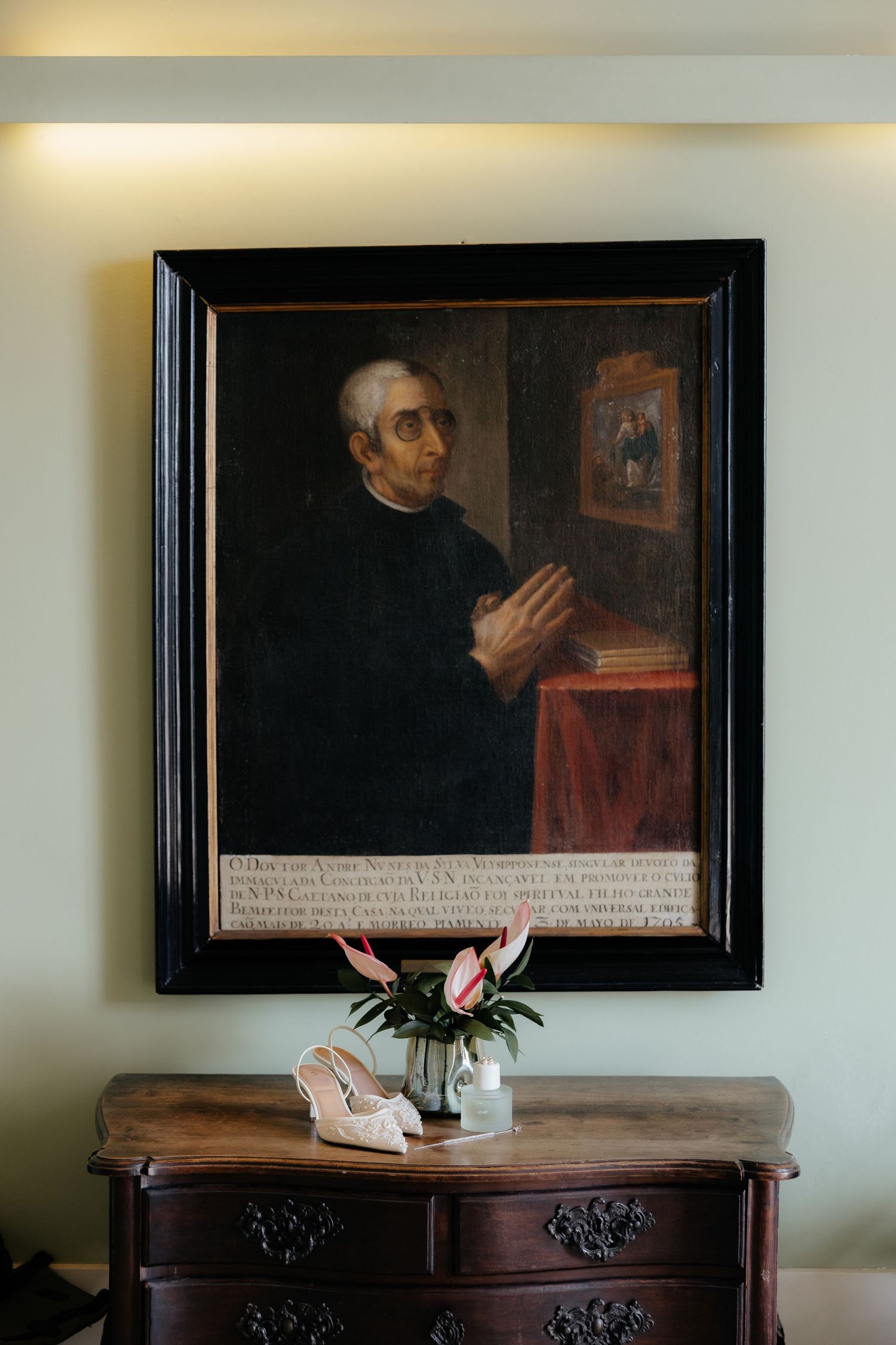 Ornate framed portrait painting hanging above a wooden dresser with bridal heels and a floral arrangement below.