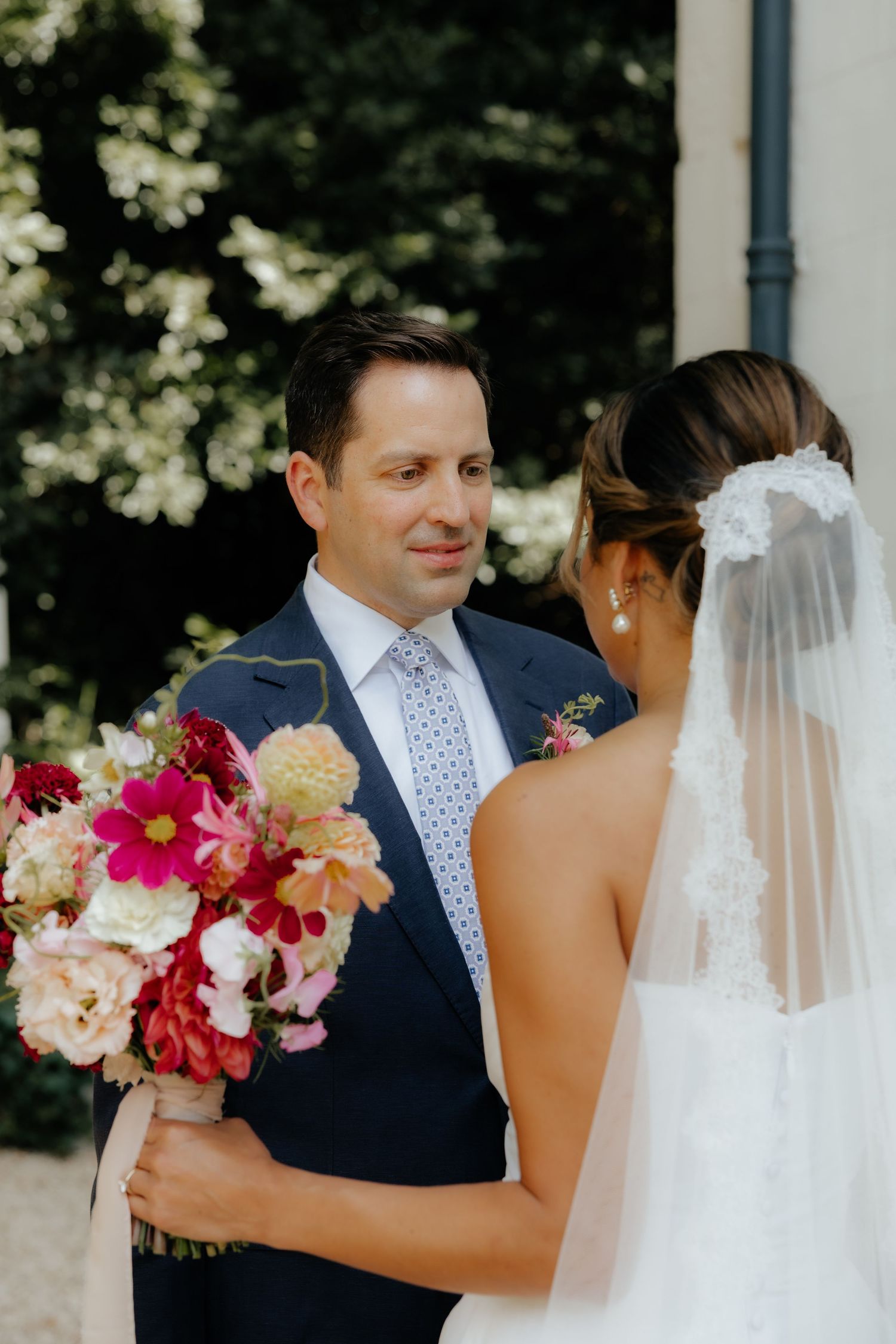 Wedding couple embraces while holding vibrant pink and peach floral bouquet against white building.