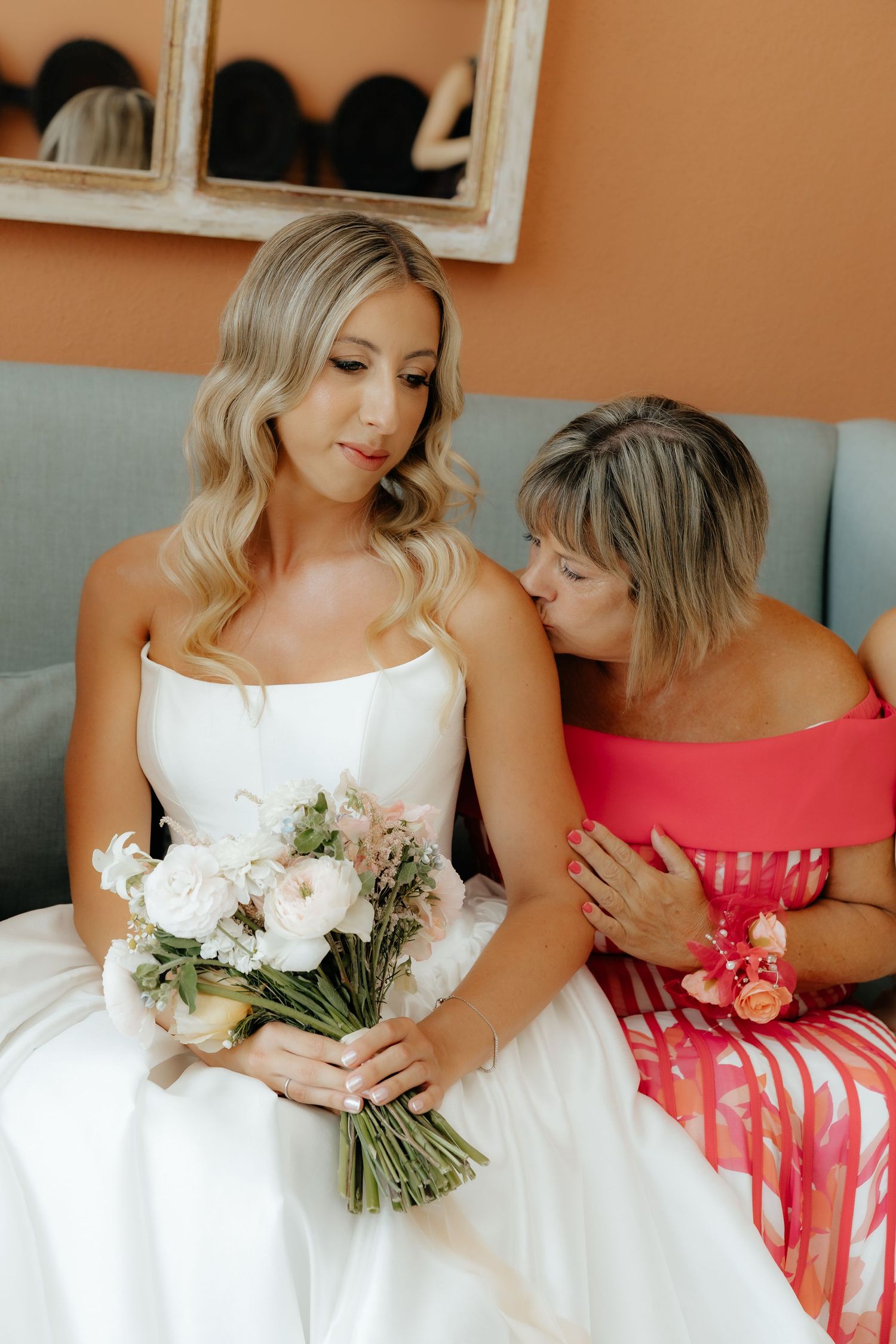 A bride in a white wedding dress holds a delicate bouquet while seated next to someone in a coral dress on a gray couch.