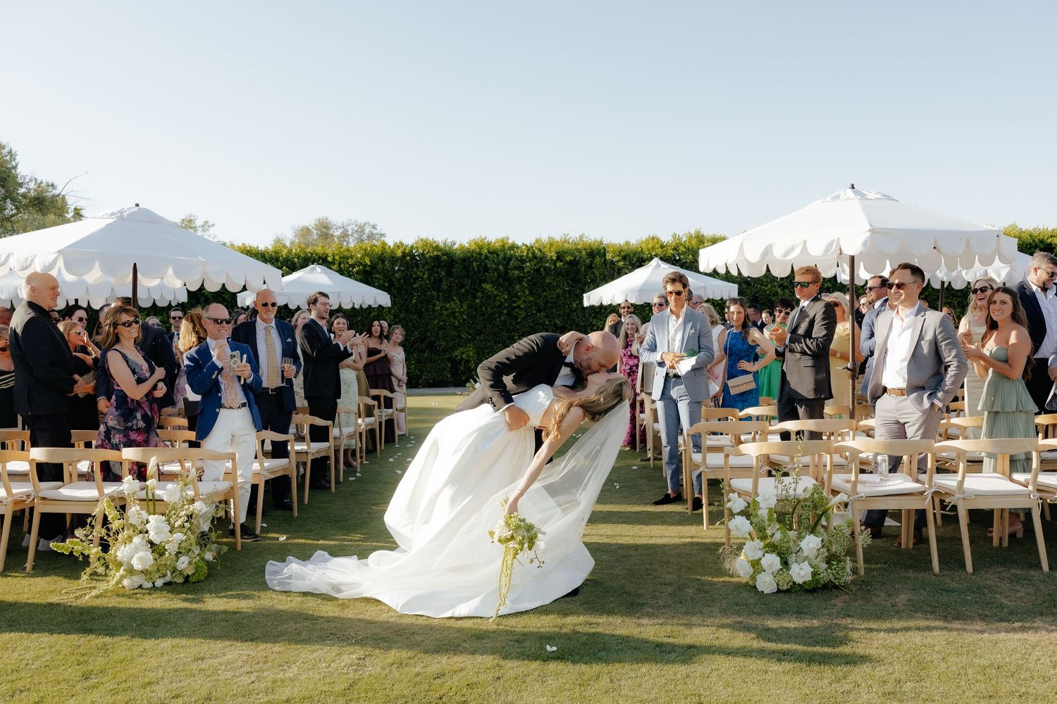 Groom dips bride in romantic kiss at outdoor wedding ceremony with seated guests applauding on both sides.