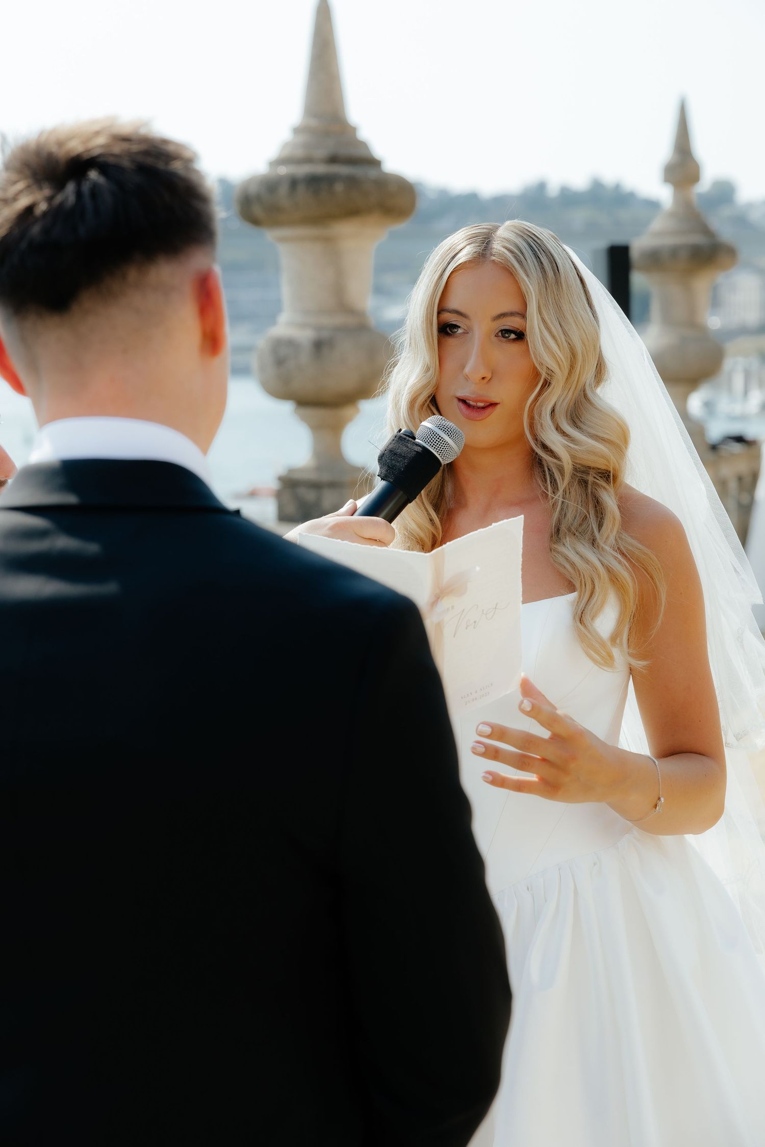 A couple exchanges wedding vows outdoors with ornate stone architectural details visible in the background.