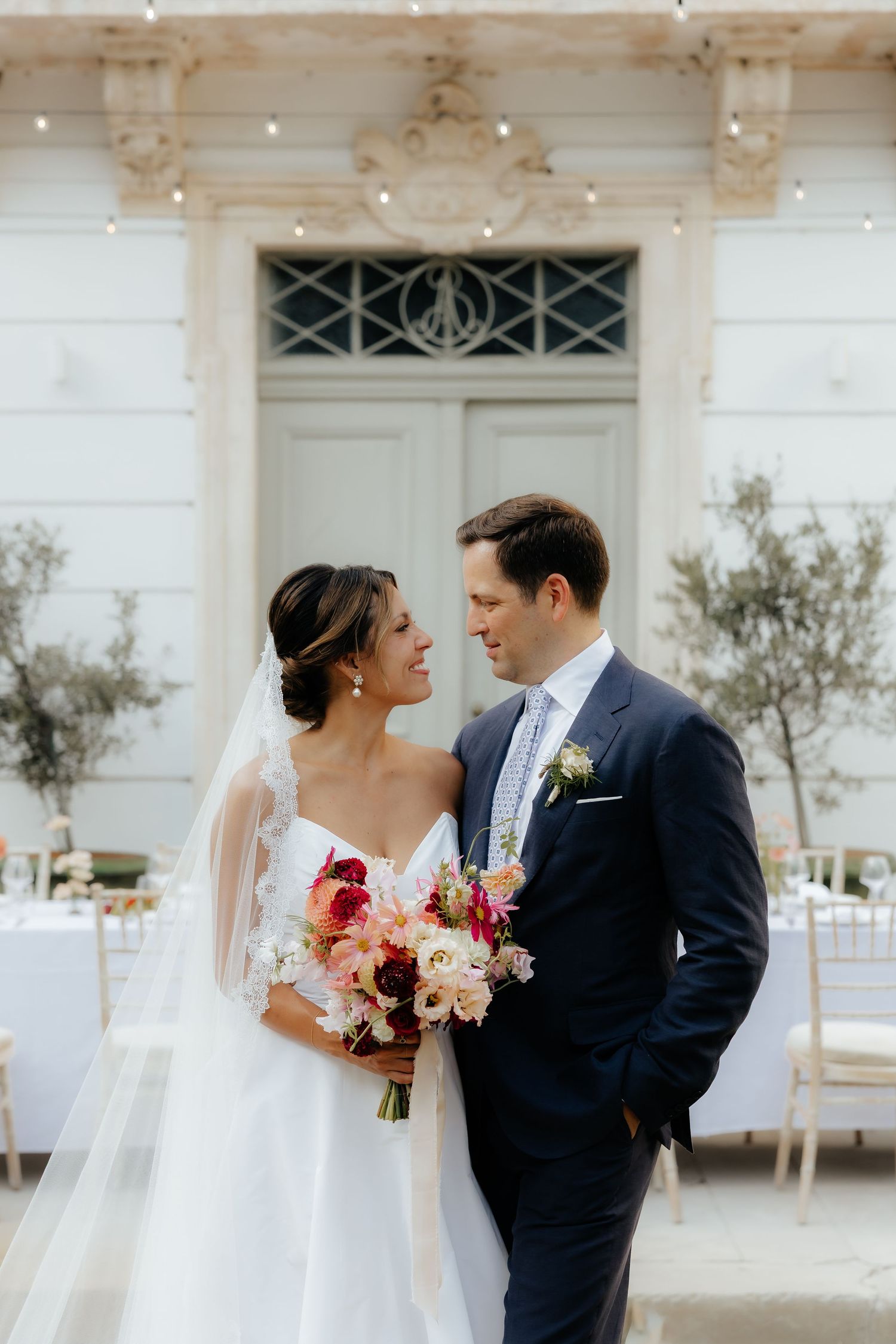 An elegant wedding scene outside a white mansion with string lights and floral decorations.