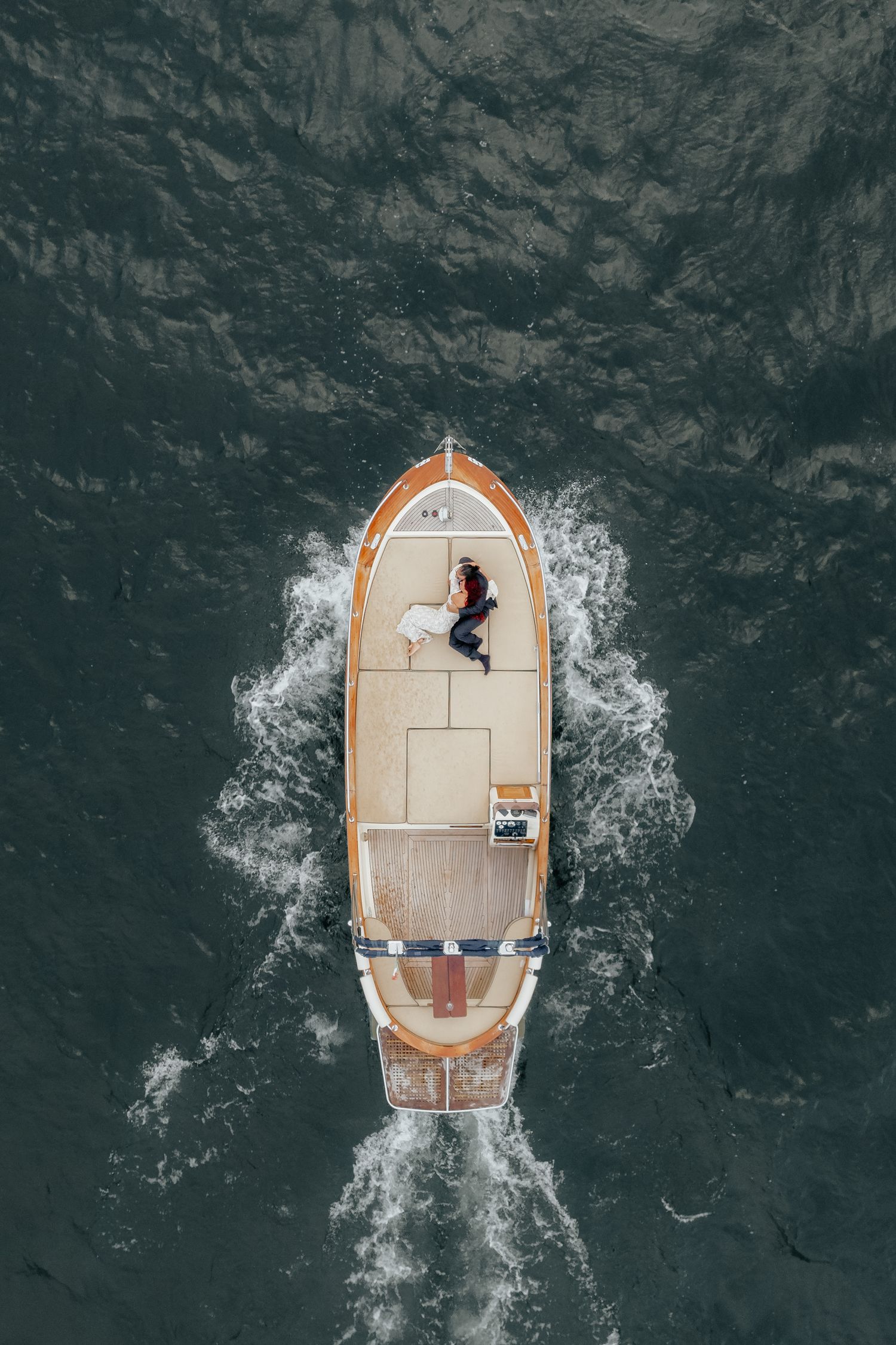 Aerial drone view of bride and groom lying on wooden boat sailing across dark water.