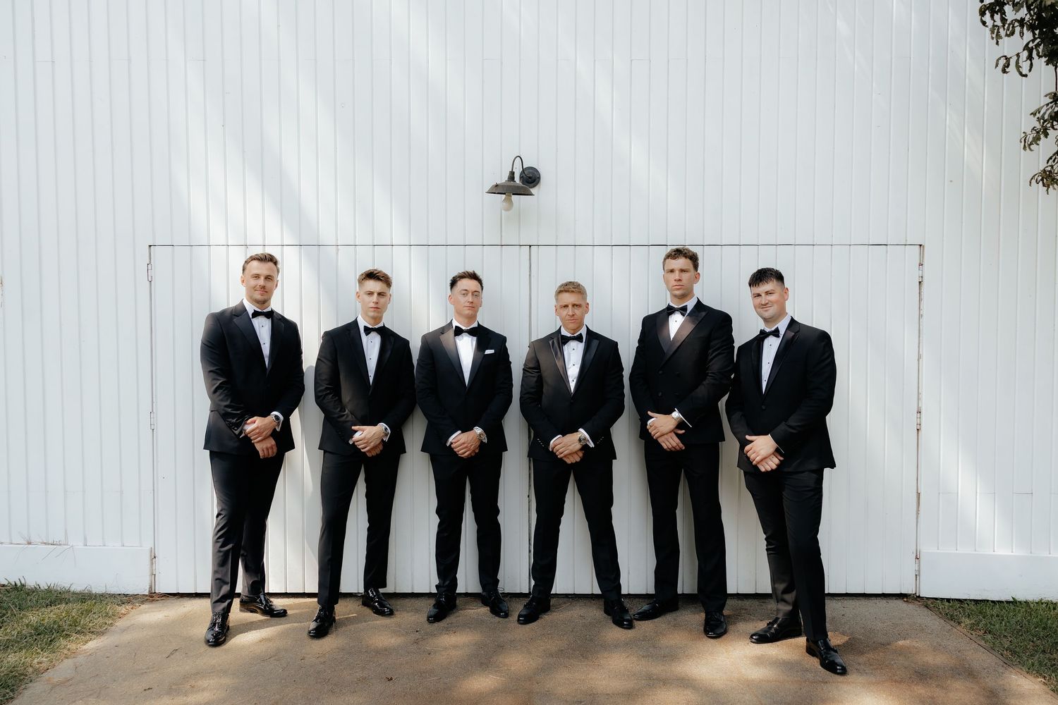 Groomsmen in black tuxedos pose in front of a white wall in a creative collage of four similar photos.