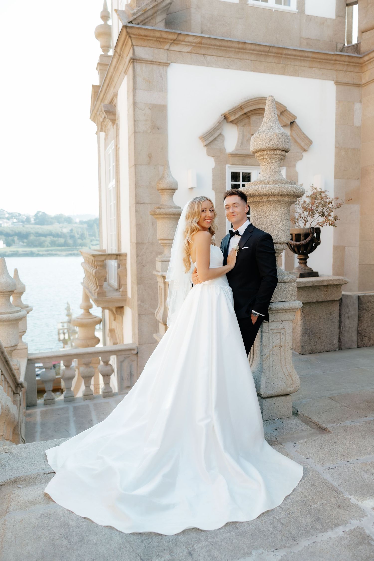 Elegant wedding couple embraces on a grand stone balcony overlooking a scenic waterfront view.