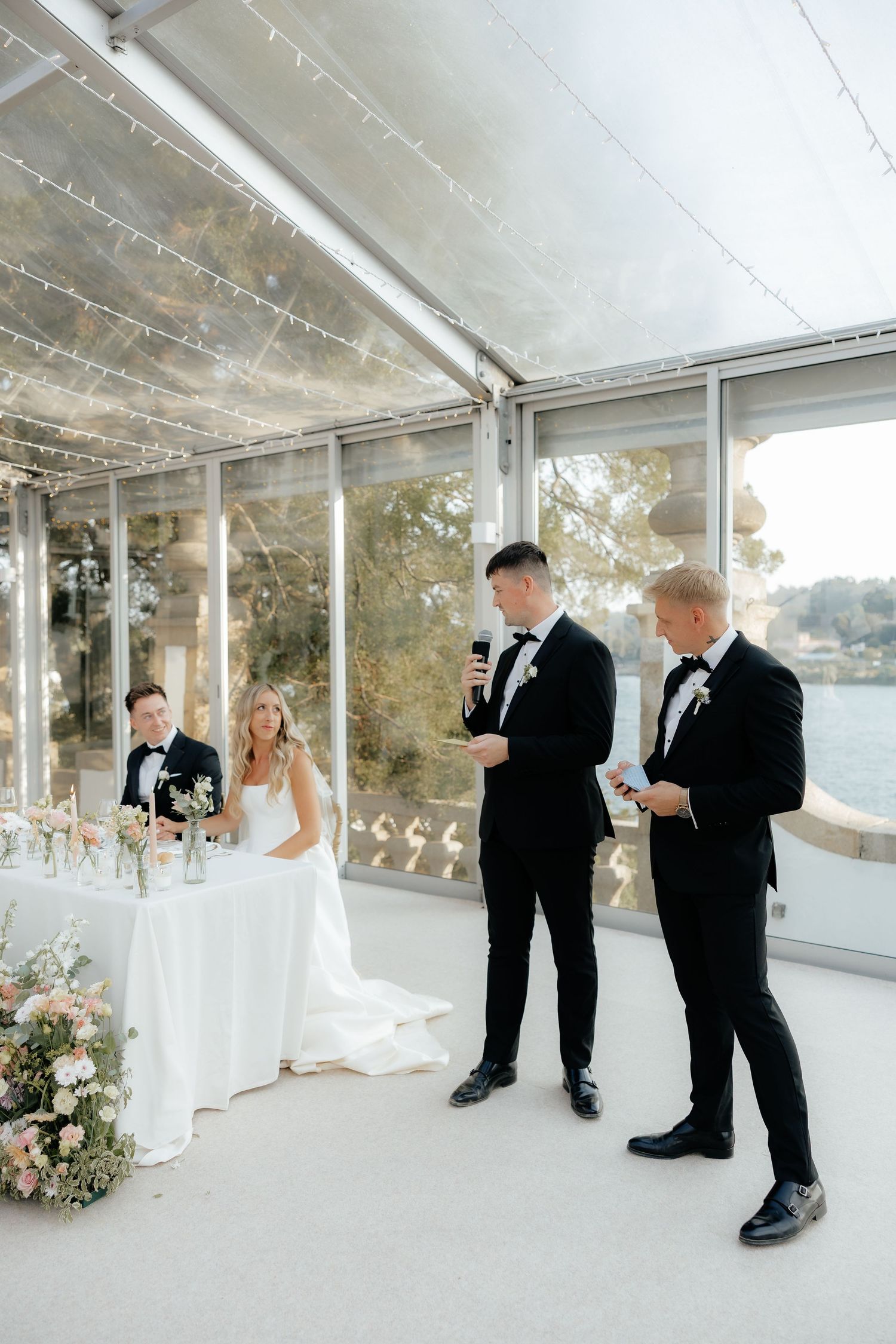 A wedding ceremony unfolds in a bright white sunroom with floor-to-ceiling windows overlooking a scenic outdoor view.