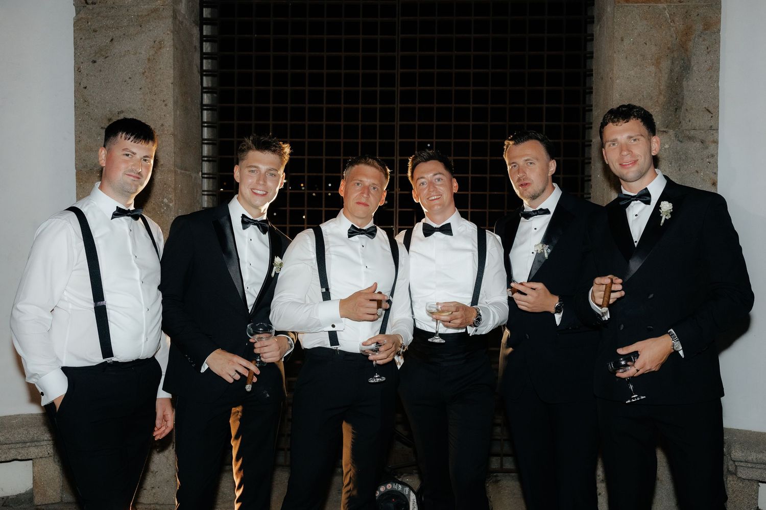 A group of men in formal black and white attire holding drinks at what appears to be a wedding celebration.