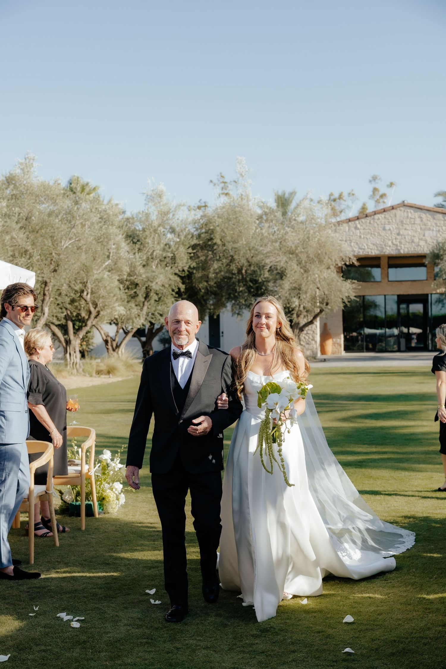 Bride in white gown holding green bouquet walks down aisle with escort at outdoor garden wedding ceremony.