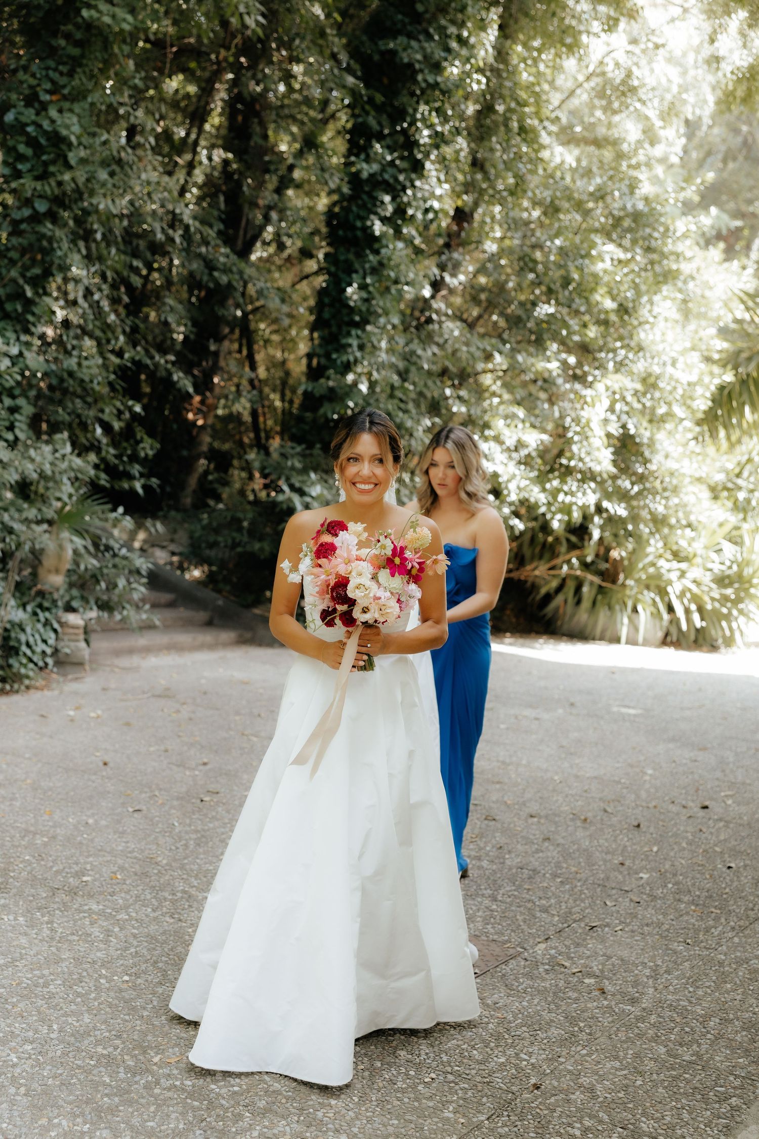 Two people in wedding attire walk together on a garden path surrounded by lush greenery.