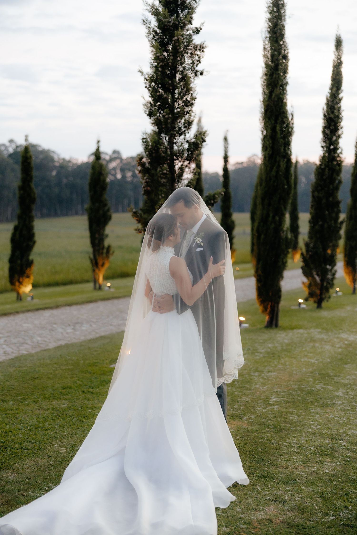 Bride and groom kiss under cathedral veil on scenic path lined with tall cypress trees at dusk.