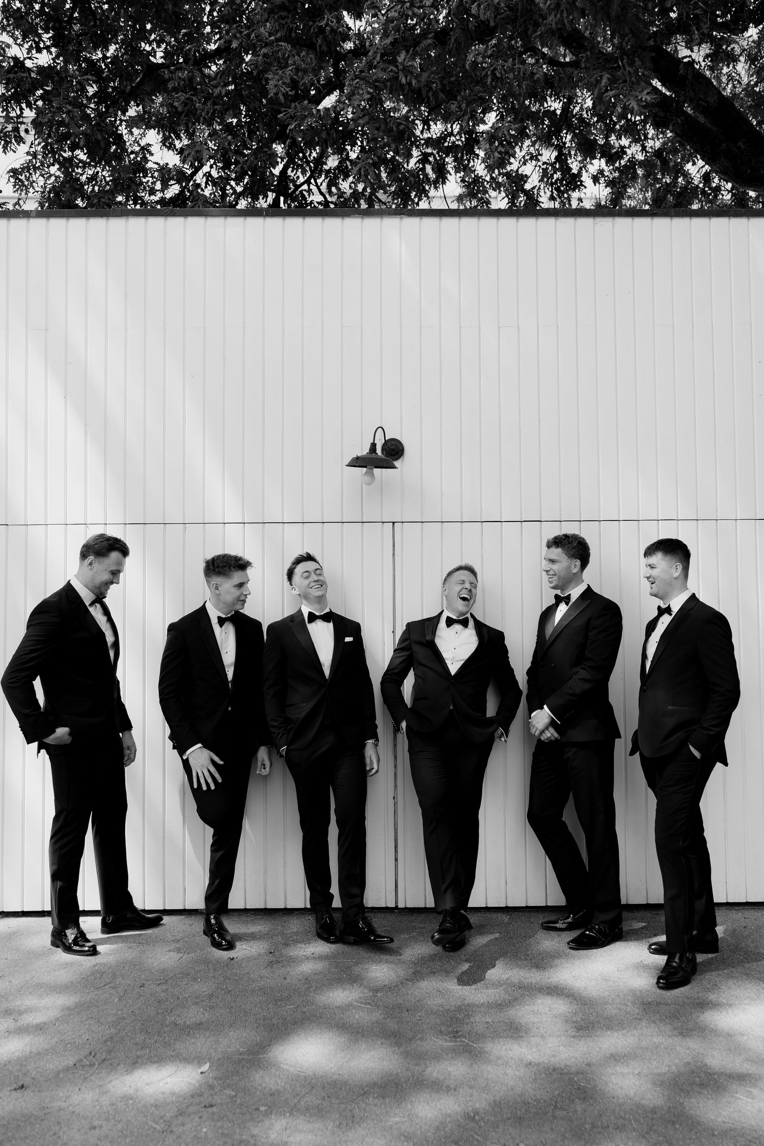 Six groomsmen in black tuxedos laughing together against a white wooden wall in a black and white wedding photo.