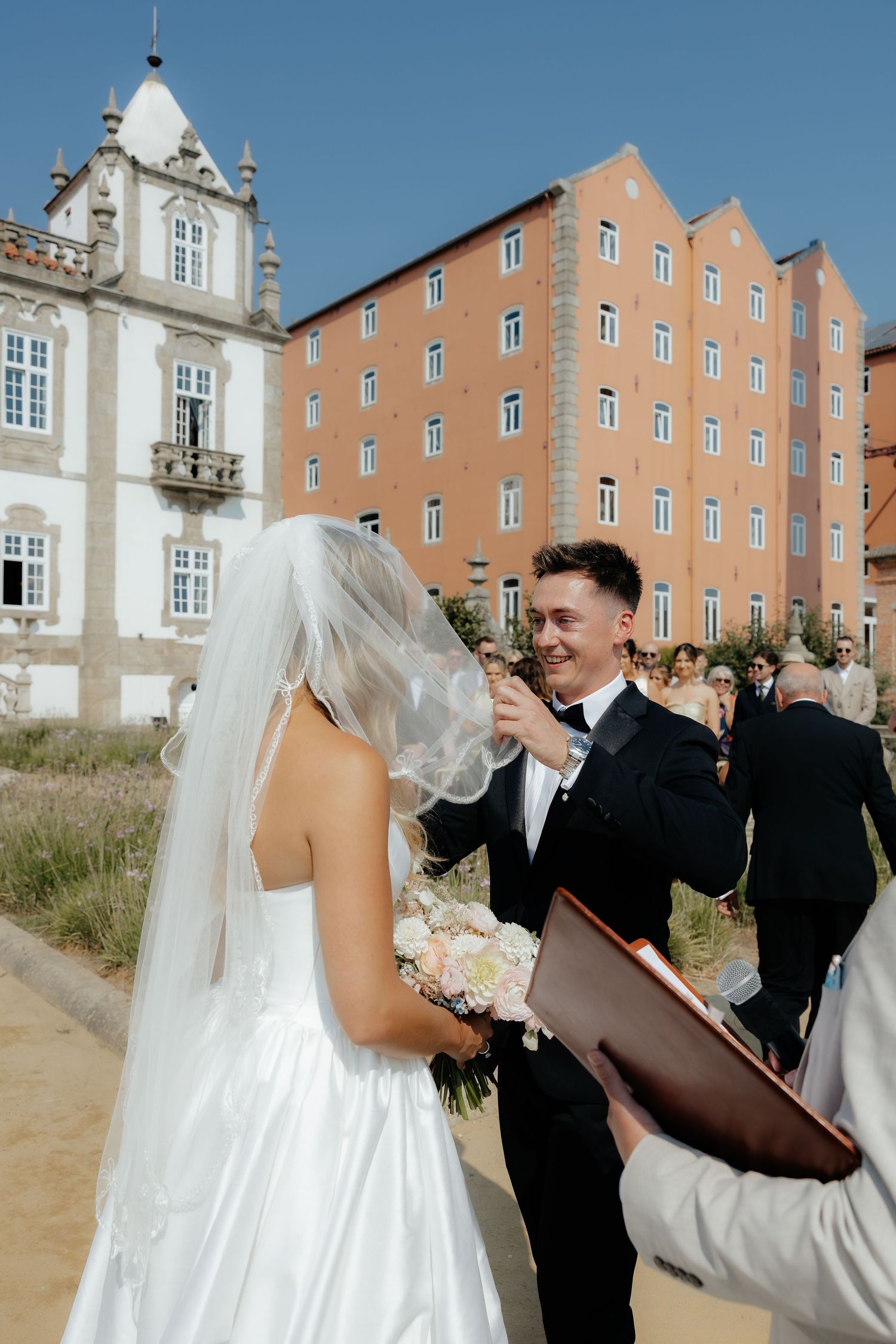 Groom smiles lifting bride's veil at outdoor wedding ceremony with historic buildings behind.
