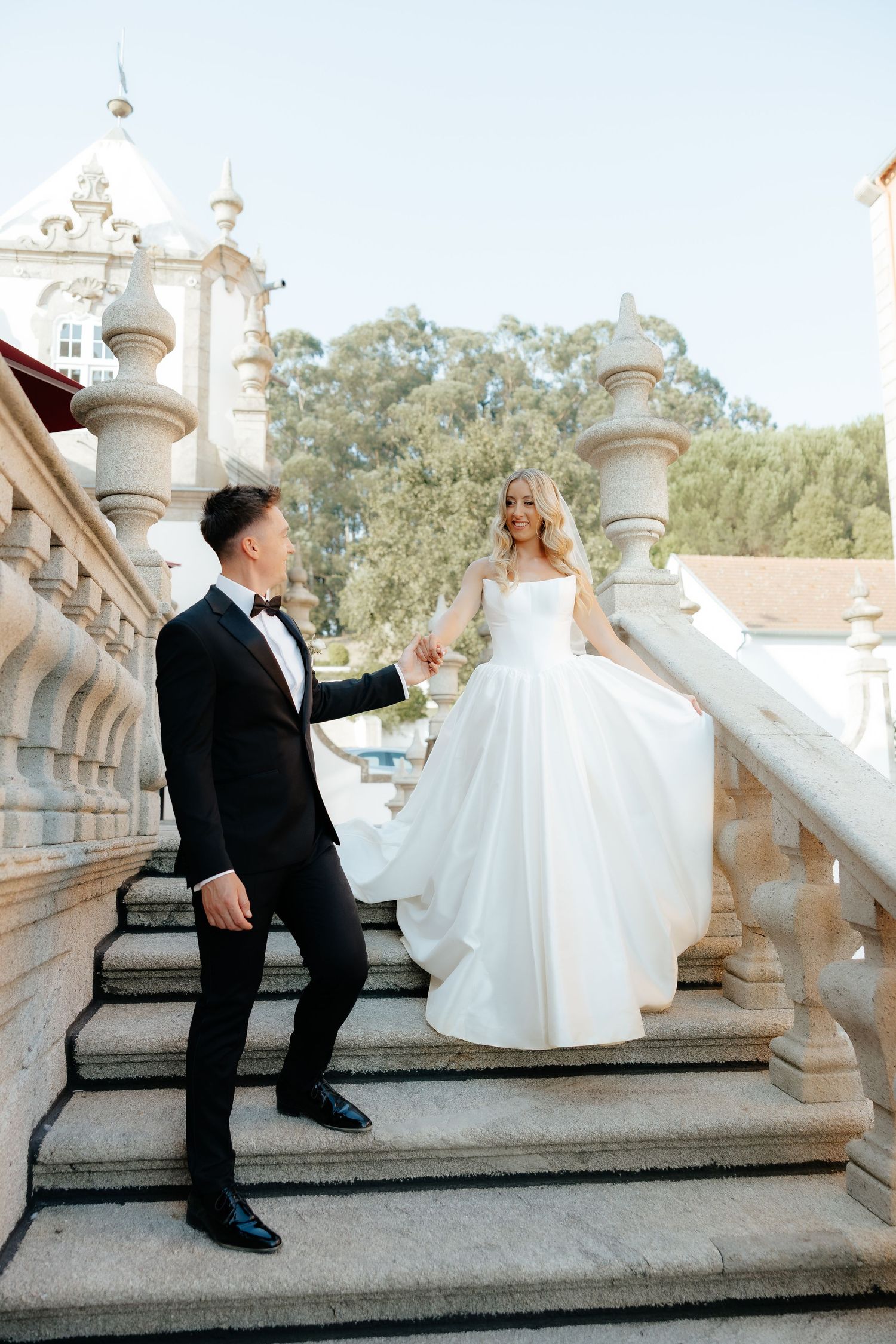 Bride and groom hold hands on grand stone staircase at elegant classical building during wedding day.