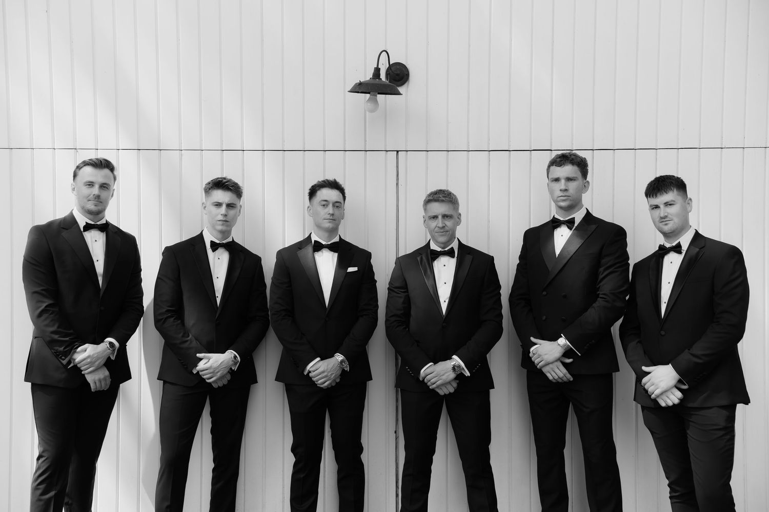 Six groomsmen in black tuxedos standing in a formal pose against a white wall in black and white.