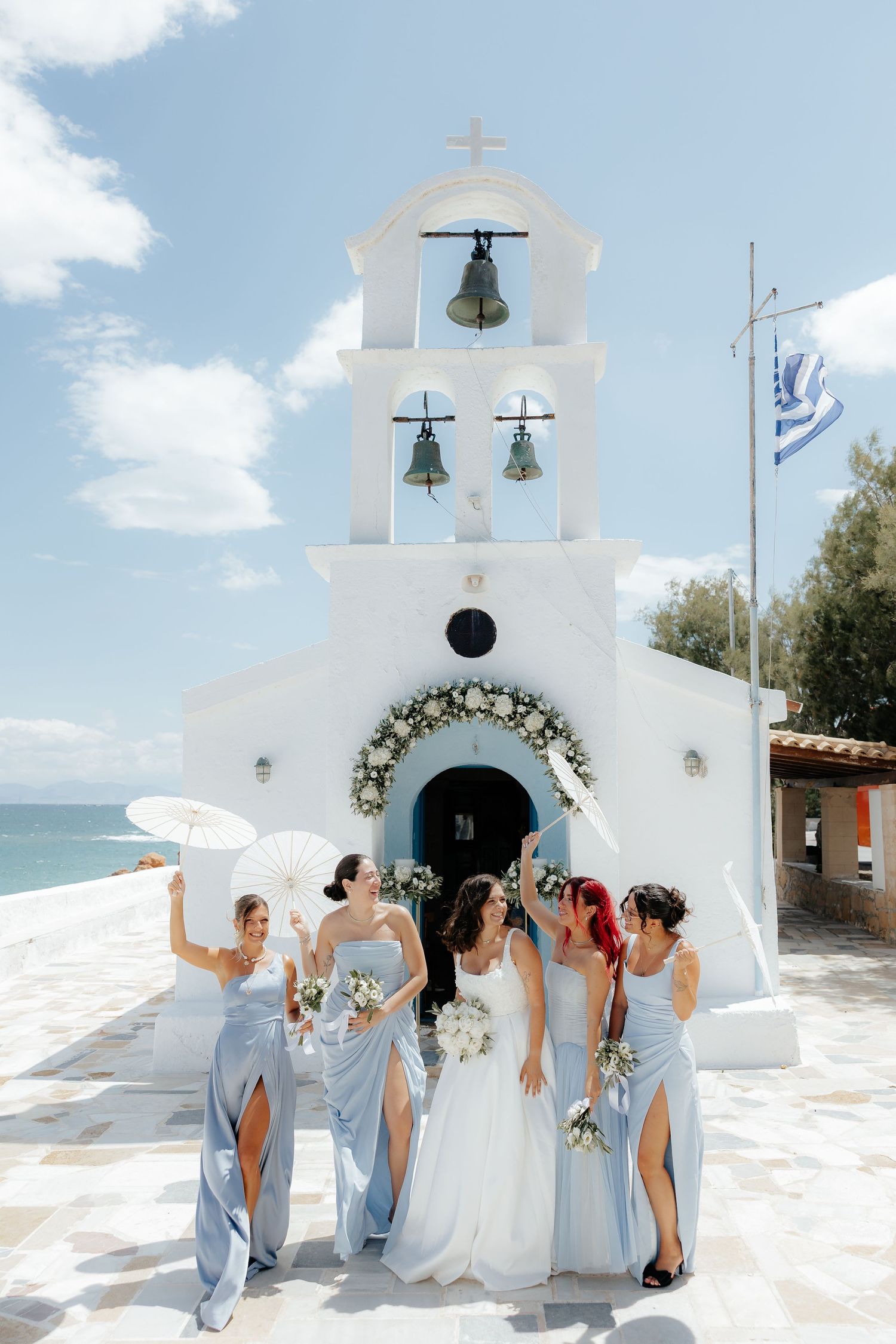 Bride and bridesmaids in blue dresses pose before a white Greek chapel with bells and Greek flag by the sea.