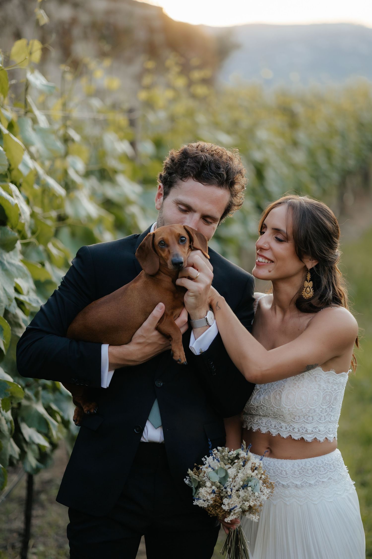 Bride and groom in vineyard holding a dachshund dog, bride in white two-piece gown holding bouquet at sunset.