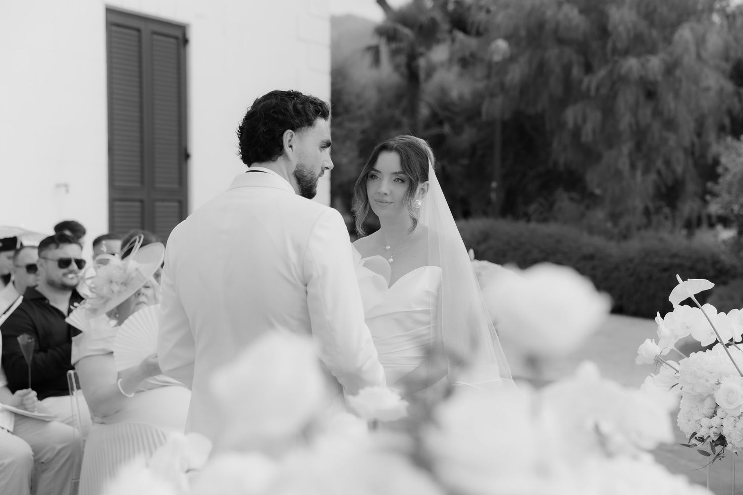 Black and white outdoor wedding ceremony with bride in veil and groom in white suit exchanging vows among guests.