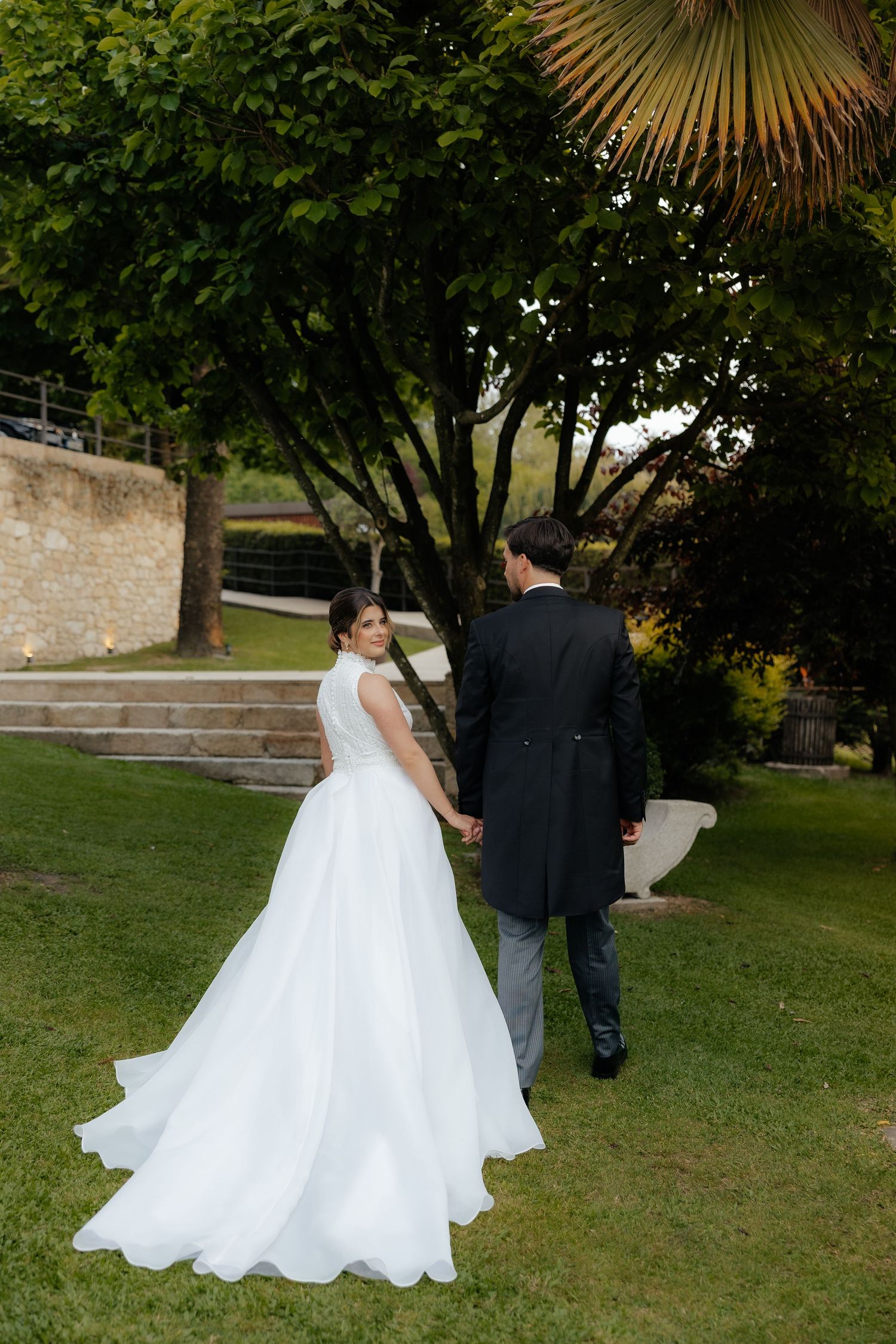 Bride in flowing white gown and groom walk hand in hand through lush green garden setting.