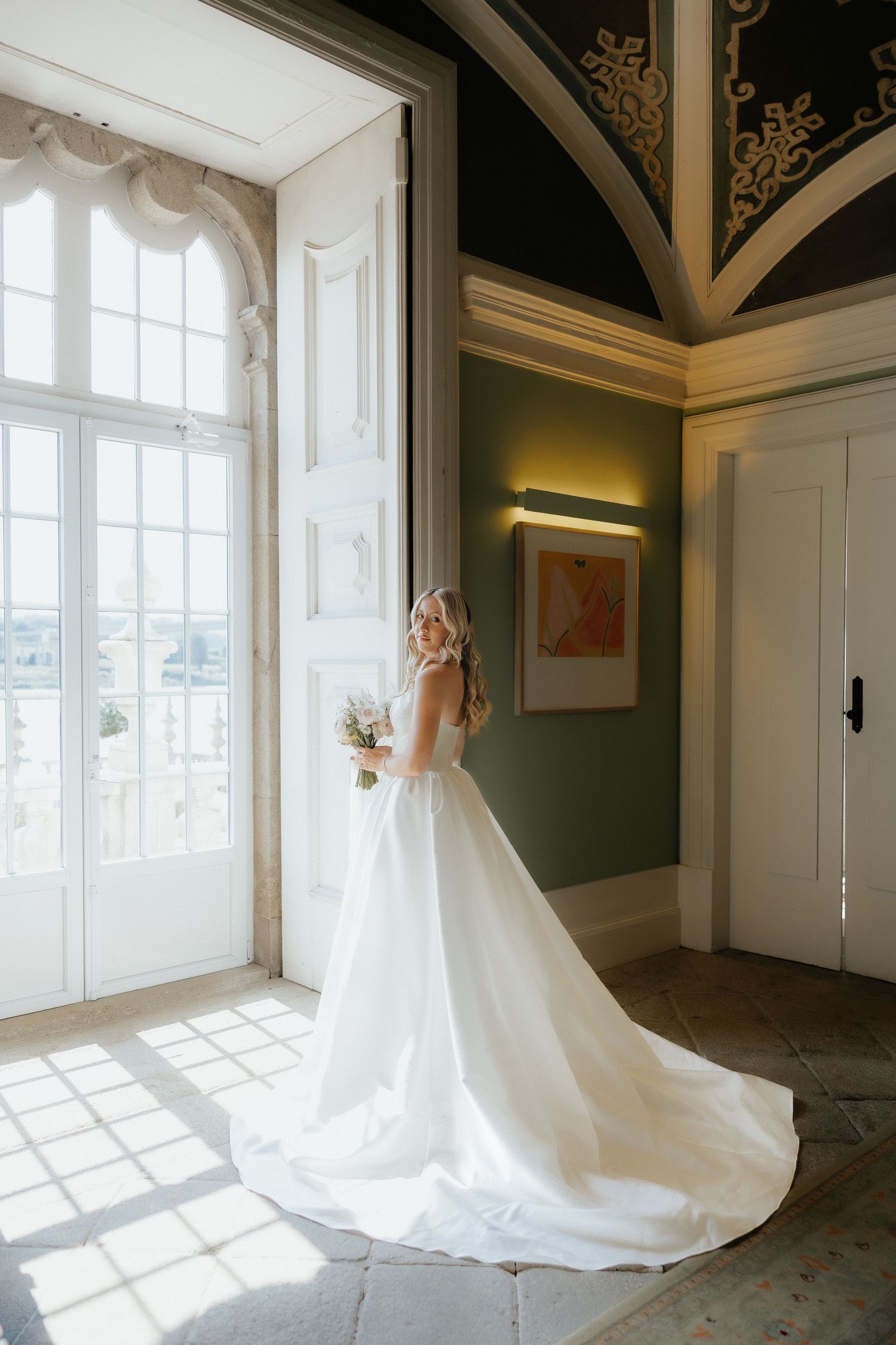 Bride in elegant white ball gown with long train stands by tall window holding bouquet in ornate venue.