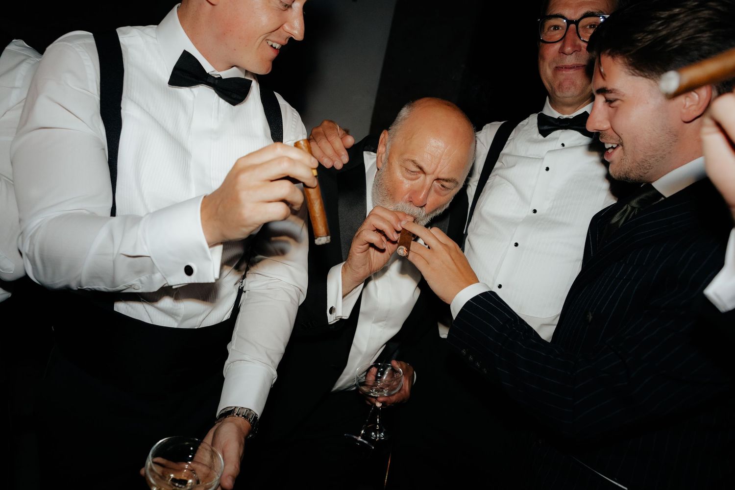 Men in tuxedos lighting cigars and enjoying drinks at a formal wedding reception celebration.