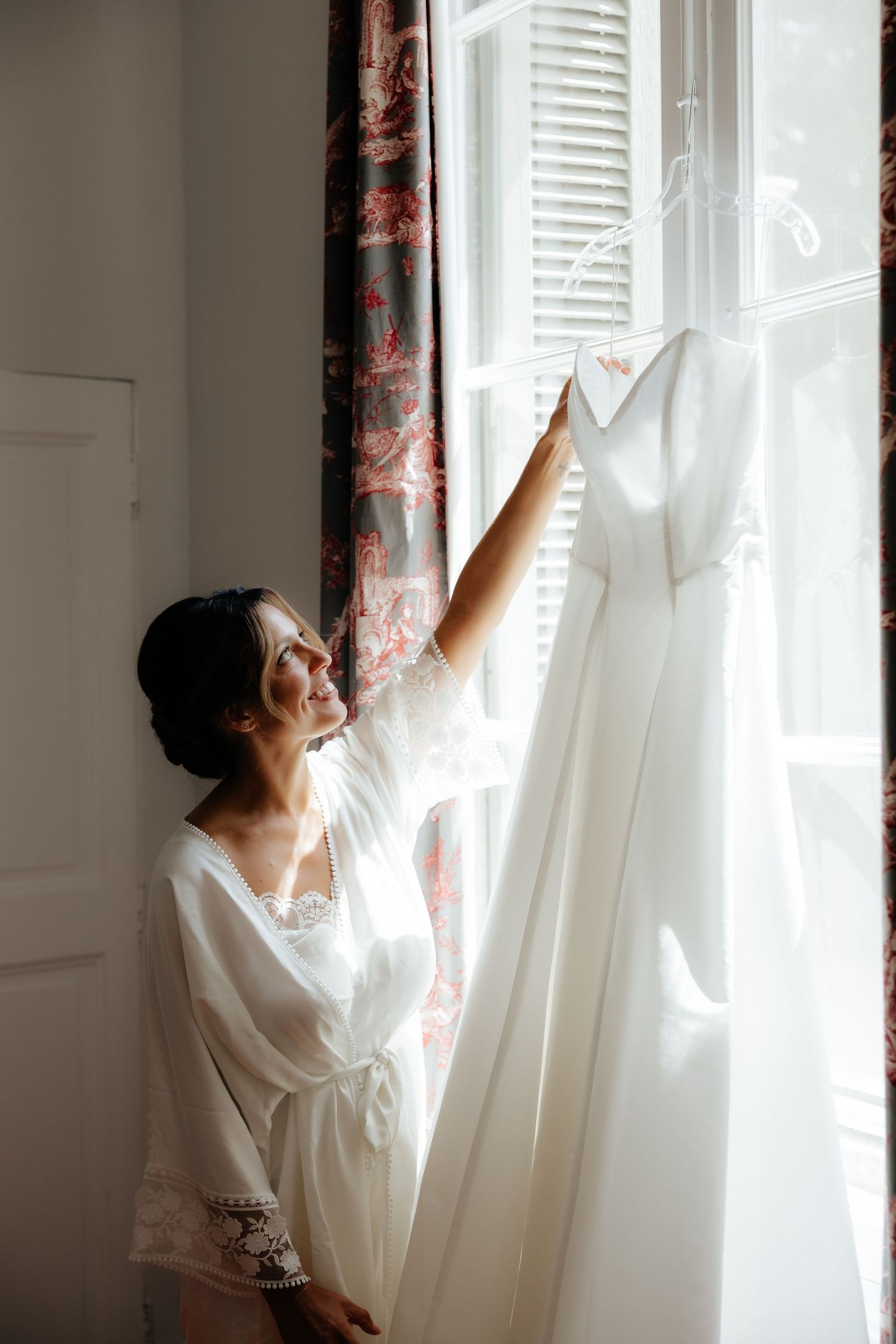 Someone in a white robe opens sheer curtains to let morning light stream through a window.