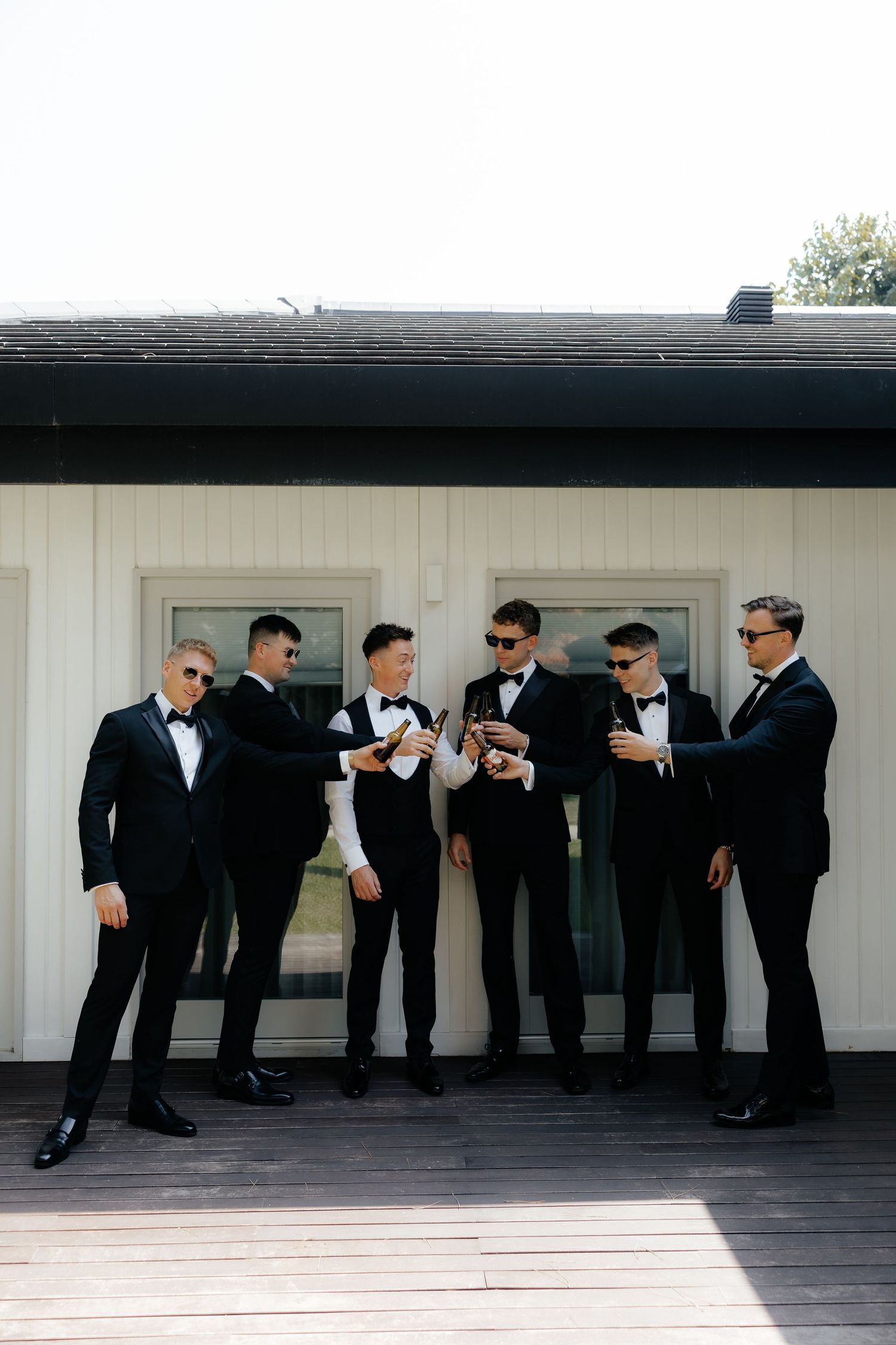 A group of groomsmen in black tuxedos and sunglasses lean forward in a domino effect pose against a white building.