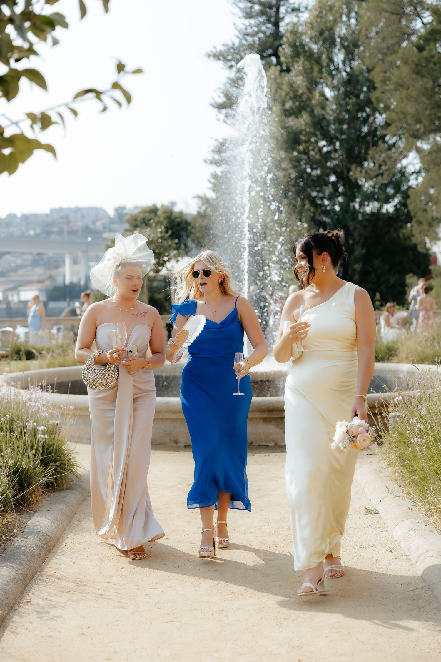 A group in formal attire walks along a sunny outdoor path with fountains spraying water in the background.