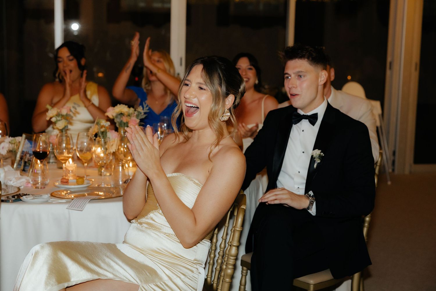 Wedding guests in formal attire laugh and celebrate during an evening reception at an elegantly decorated venue.