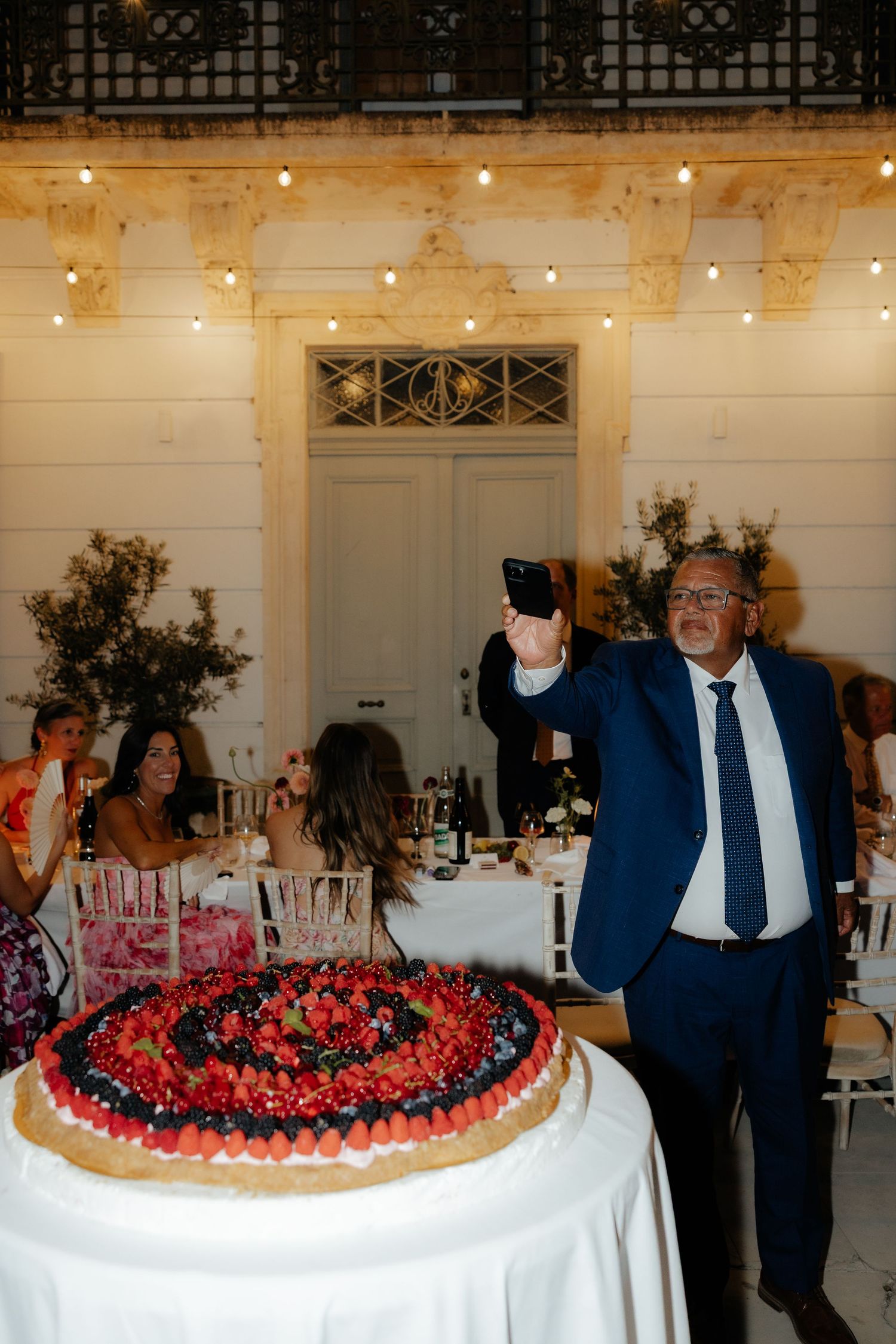 A large round cake decorated with fresh berries is being presented at an elegant indoor reception venue.