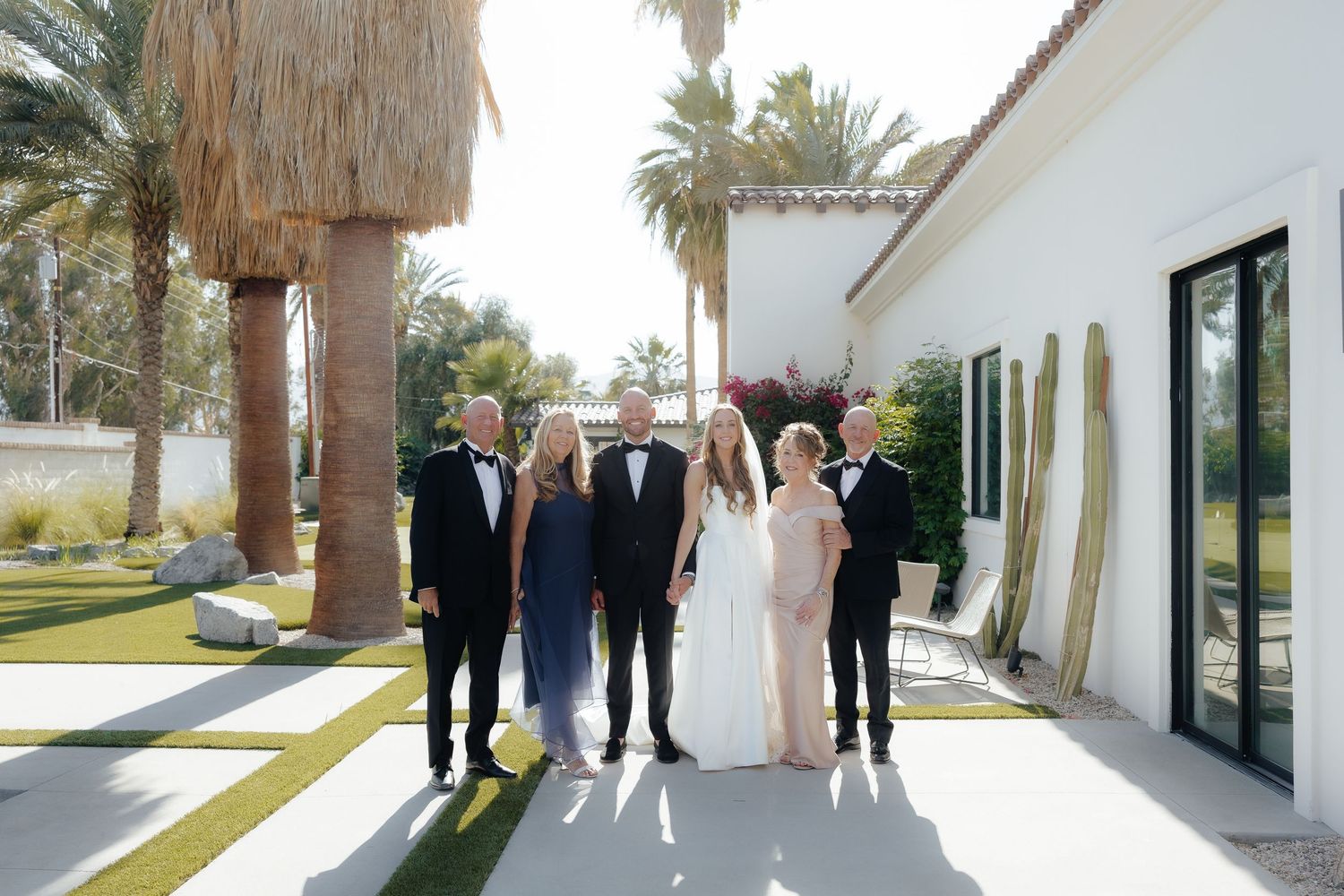 Six wedding guests posing together outside a white villa surrounded by palm trees.