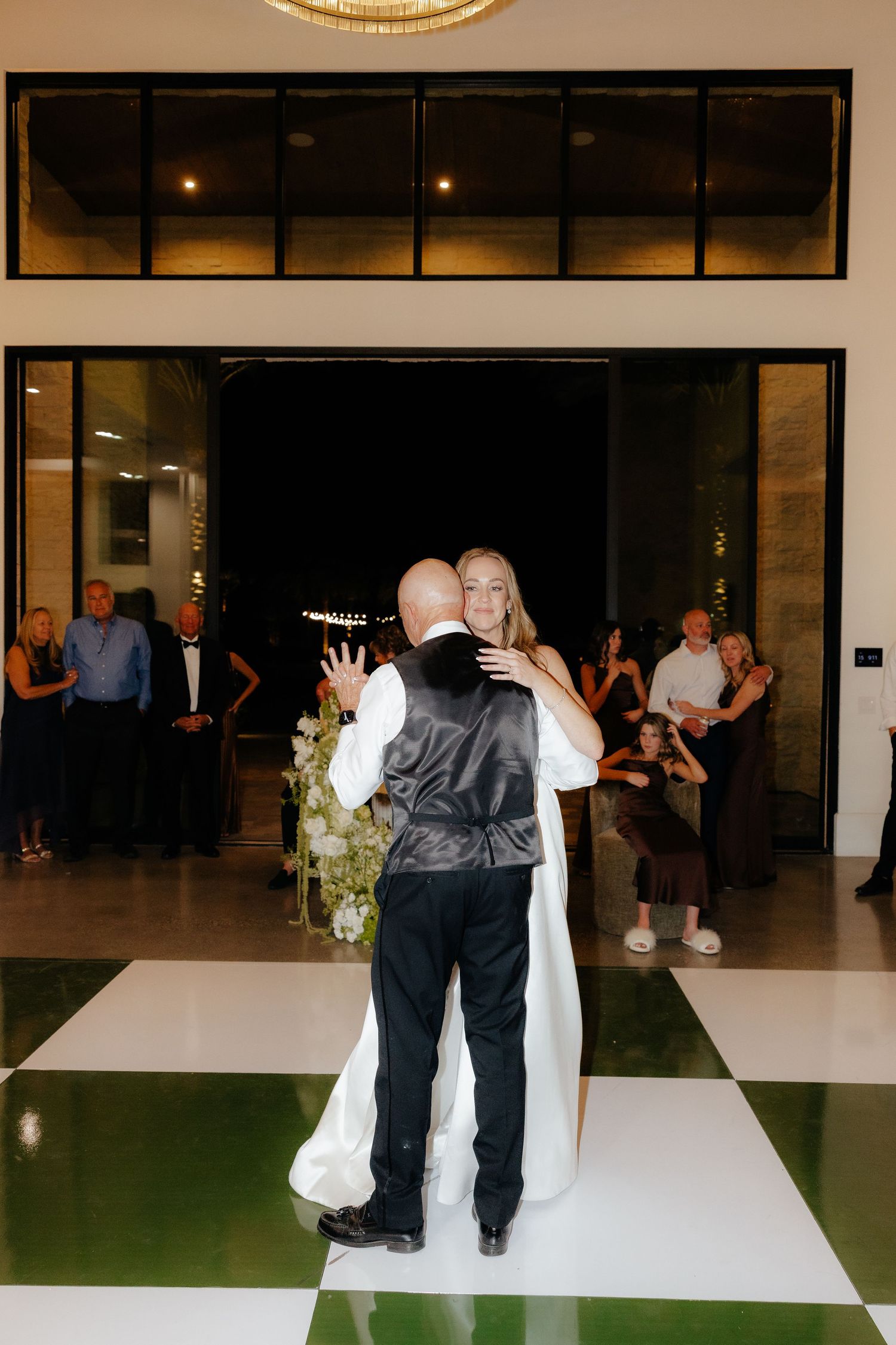 Bride and groom share their first dance on a green and white checkered floor while wedding guests look on.