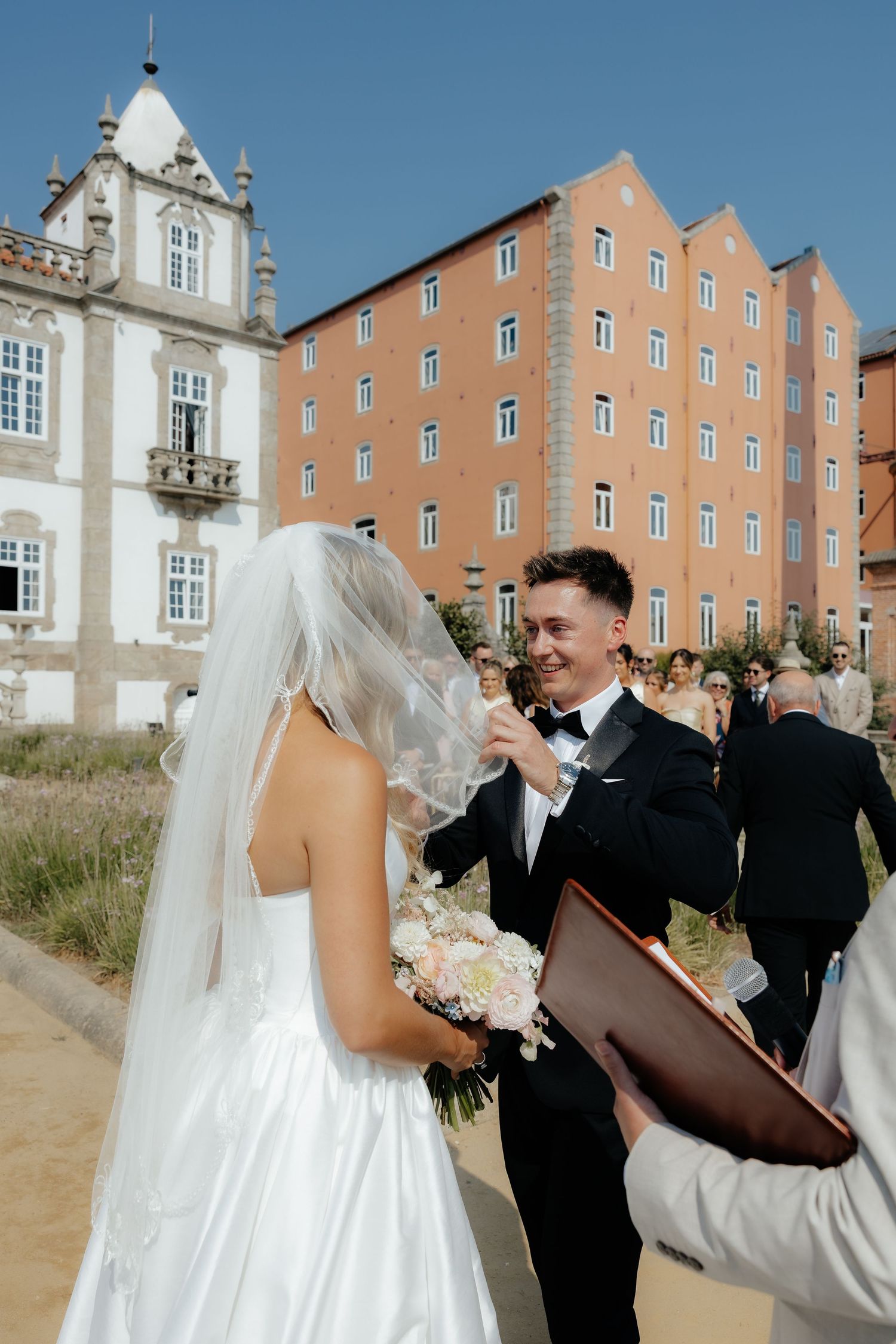 A bride and groom share a joyful moment together outside a grand historic building with peach-colored walls.