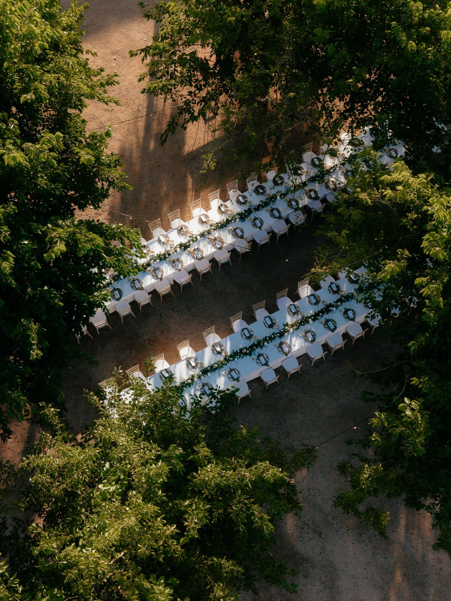 Aerial drone view of elegant outdoor wedding reception tables set among lush green trees in dappled sunlight.