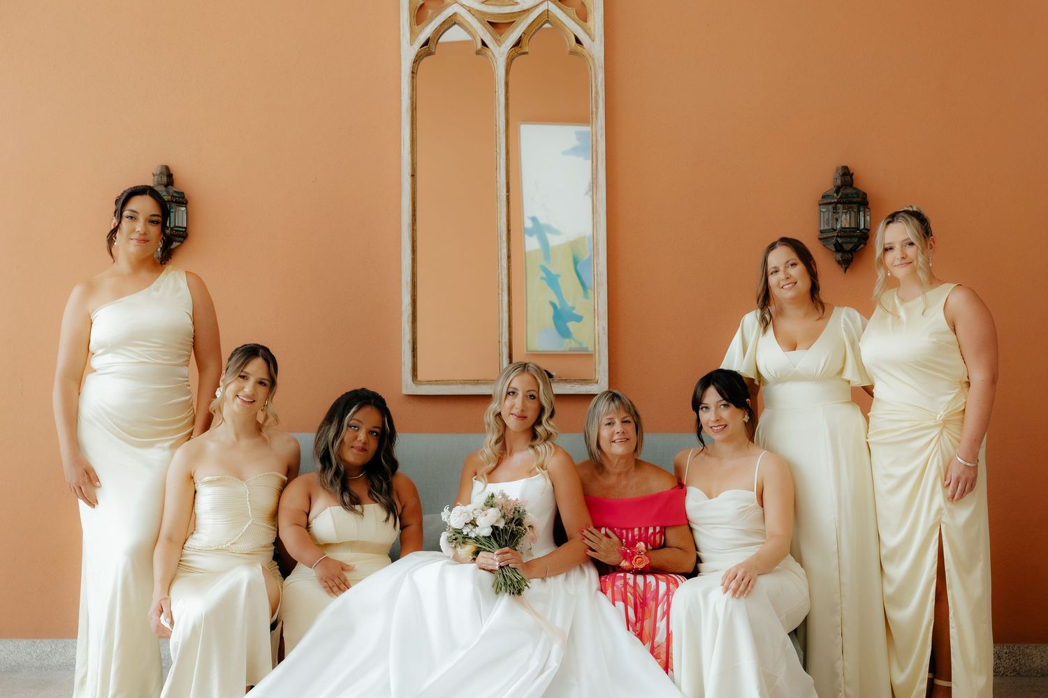 A row of bridesmaids in cream-colored dresses pose together against a peach-colored wall at a wedding.