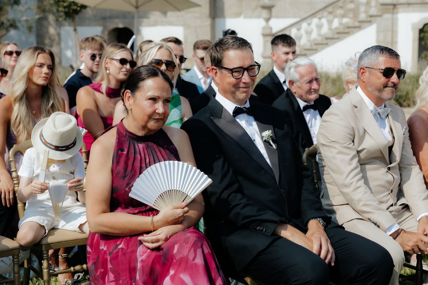 Wedding guests sit in rows of white chairs during an outdoor ceremony on a sunny day.